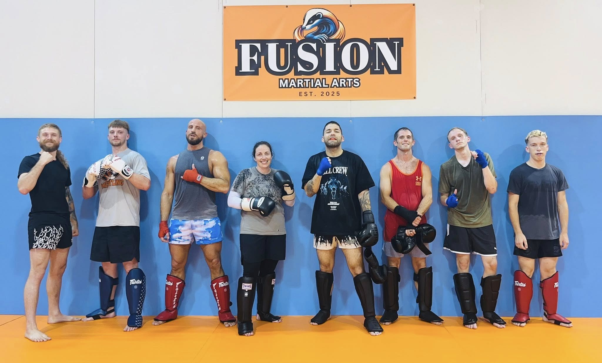 A group of eight martial artists standing in a gymnasium in front of a sign that reads "Fusion Martial Arts." They are wearing boxing gloves and protective gear, smiling and posing for the photo.