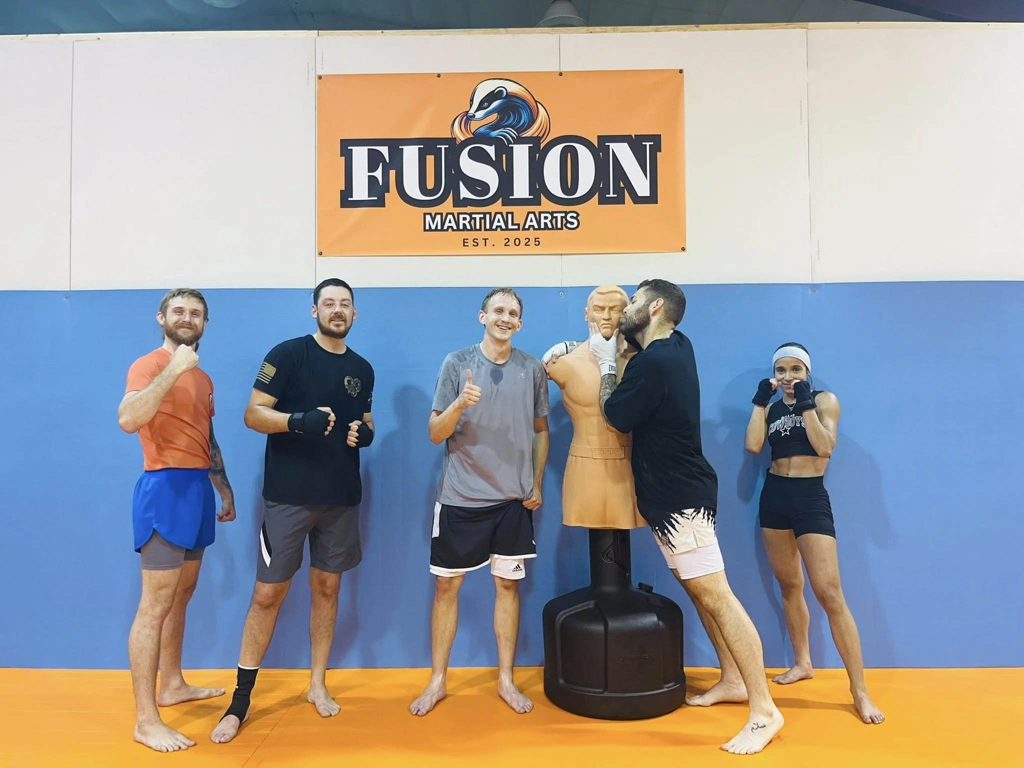 Group of six martial artists in training gear is standing in a gym with blue and white walls and an orange mat, posing in front of a Fusion Martial Arts banner.