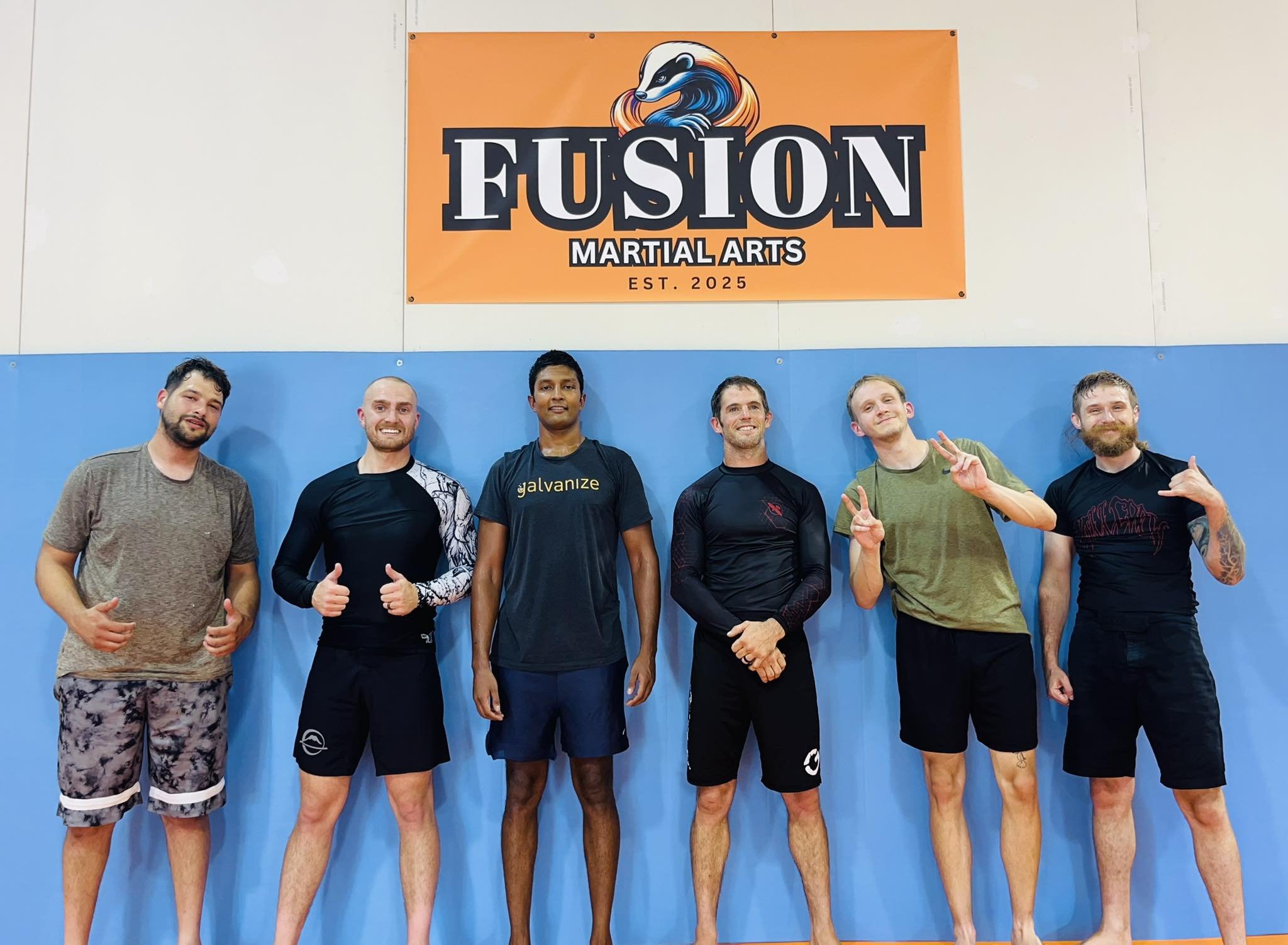 Six men posing in a martial arts gym, standing in front of a blue wall and an orange banner that reads 'Fusion Martial Arts EST. 2025' with a honey badger logo.