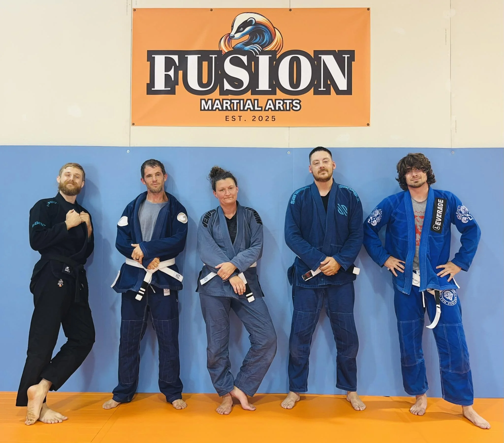 Five people dressed in Brazilian Jiu-Jitsu gi uniforms standing in a row on an orange mat in Fusion Martial Arts gym, posing for a group photo.