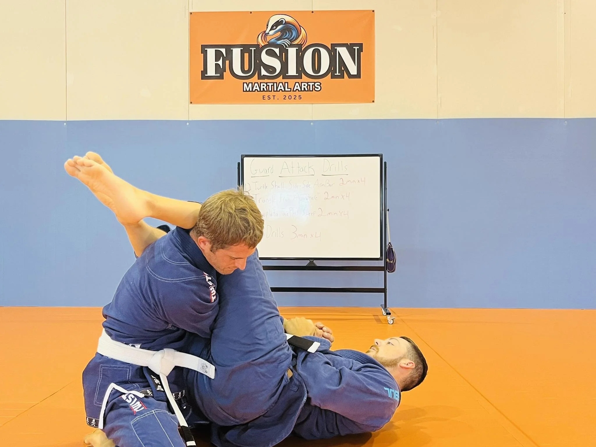 Two men practicing Brazilian Jiu-Jitsu on an orange mat at Fusion Martial Arts. One man is on the ground in a blue gi, and the other man is on top in a navy gi, controlling him. A whiteboard with handwritten instructions is in the background, and a F