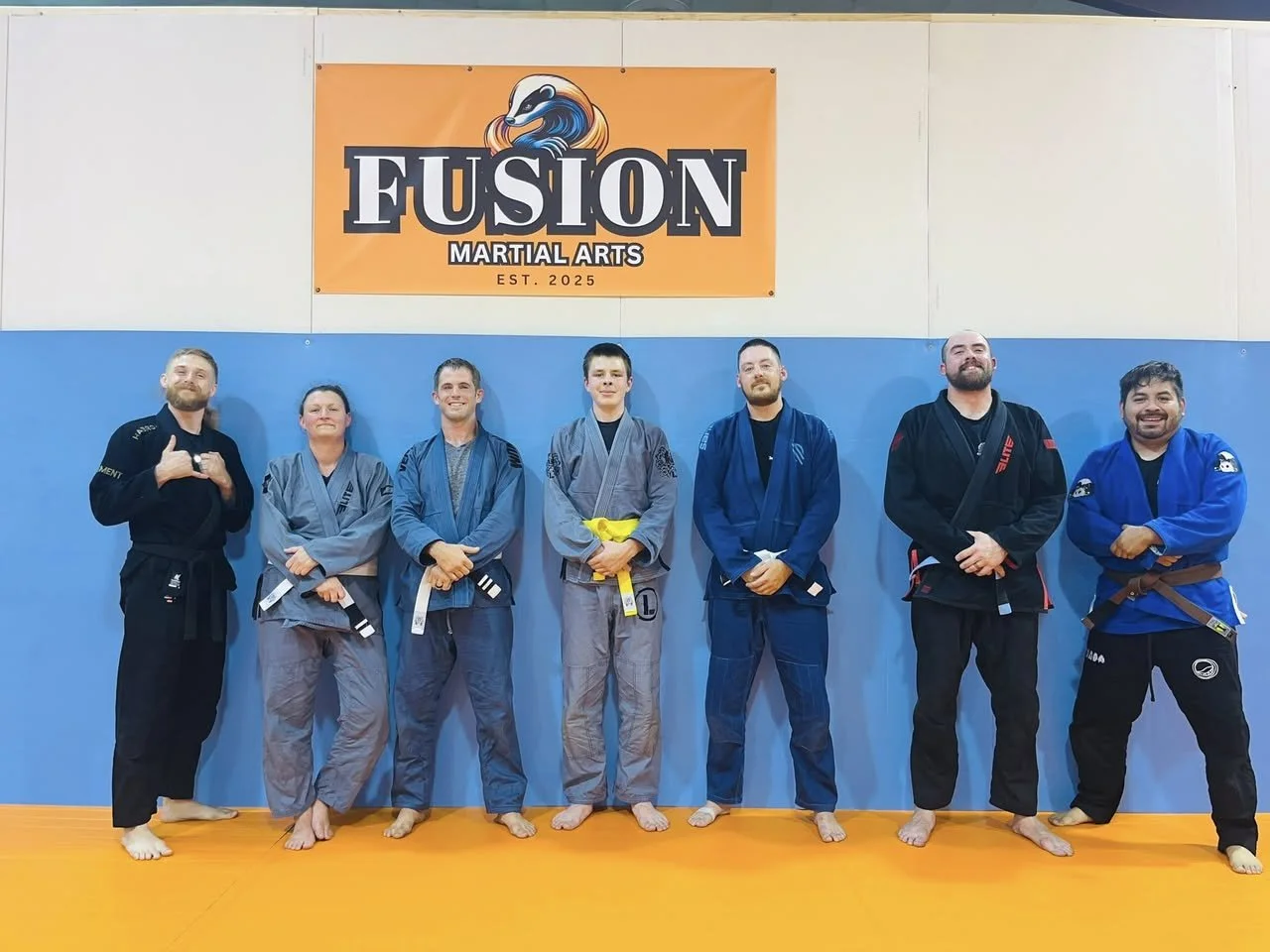 Seven people in martial arts gi uniforms standing in a row on an orange mat inside a dojo, with a Fusion Martial Arts sign on the wall behind them.