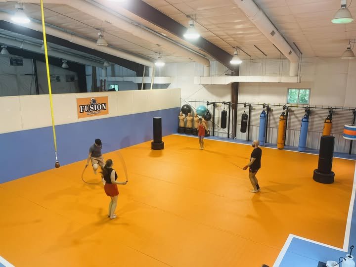 Inside a martial arts, jiu-jitsu gym with an orange mat floor, three people are jumping rope, and one person is holding a staff. Gym equipment like punching bags and exercise balls are visible in the background.
