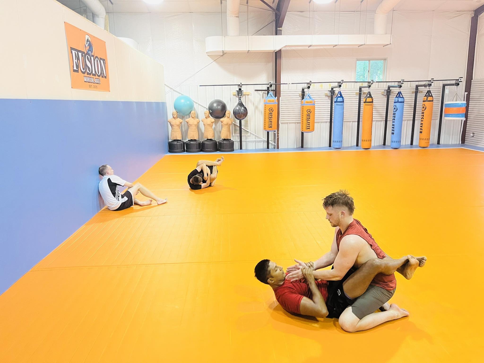 Two men are practicing Brazilian Jiu-Jitsu on an orange mat, with one man in red training gear on top of another man in black. In the background, two children and one adult are sitting on the edge of the mat. The gym has blue and white walls, with tr