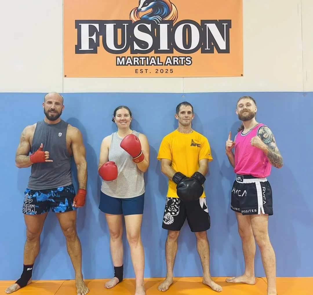 Four individuals in martial arts training gear standing in a gym, smiling, with a large sign in the background that reads 'Fusion Martial Arts'.