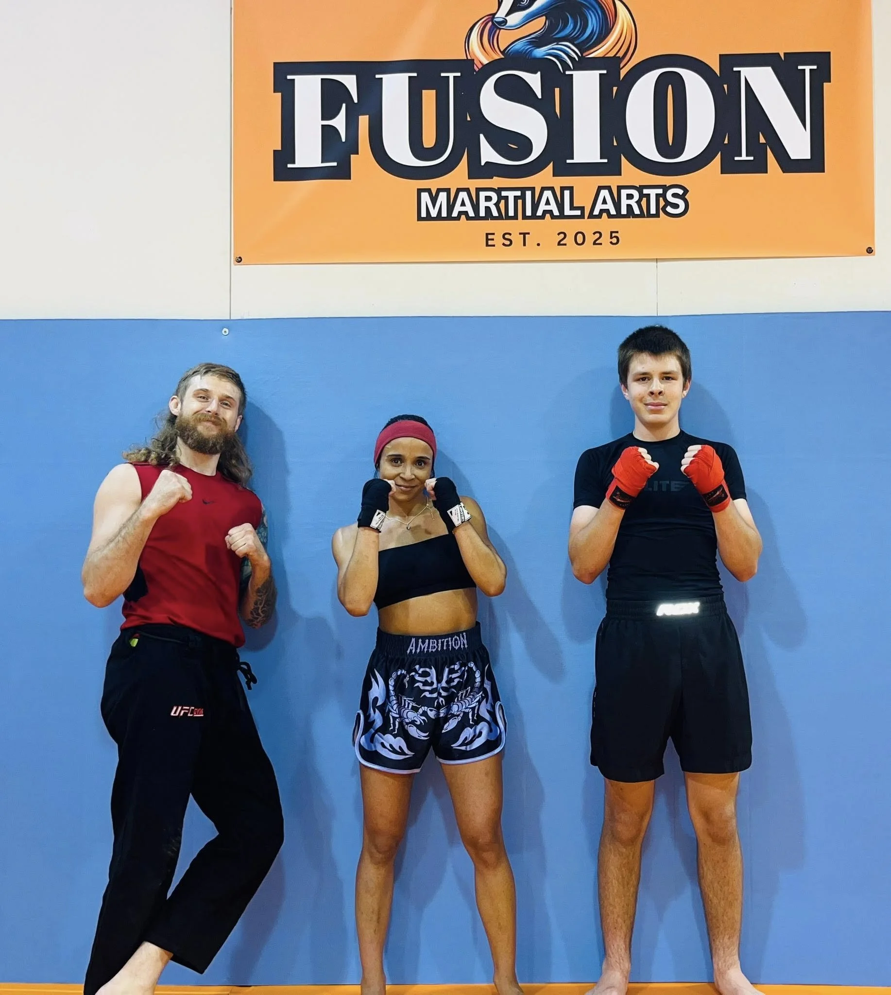 Three martial artists standing in fighting stance, posing inside a gym with a banner that reads 'Fusion Martial Arts' and 'Est. 2025' behind them.