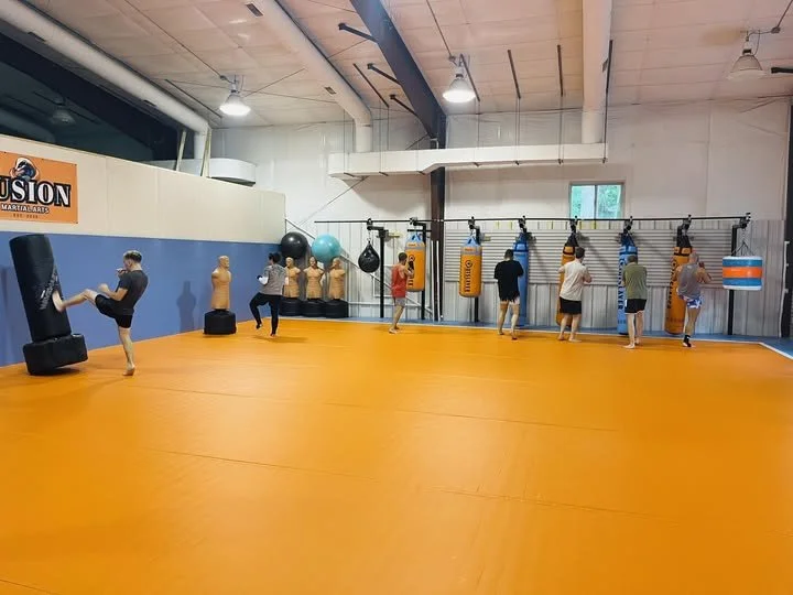 People practicing kickboxing with punching bags and training equipment in a gym with orange flooring and a white and blue wall.