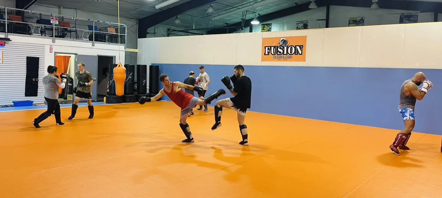 People practicing martial arts in a gym with orange flooring, including fighters wearing gloves and pads for training.