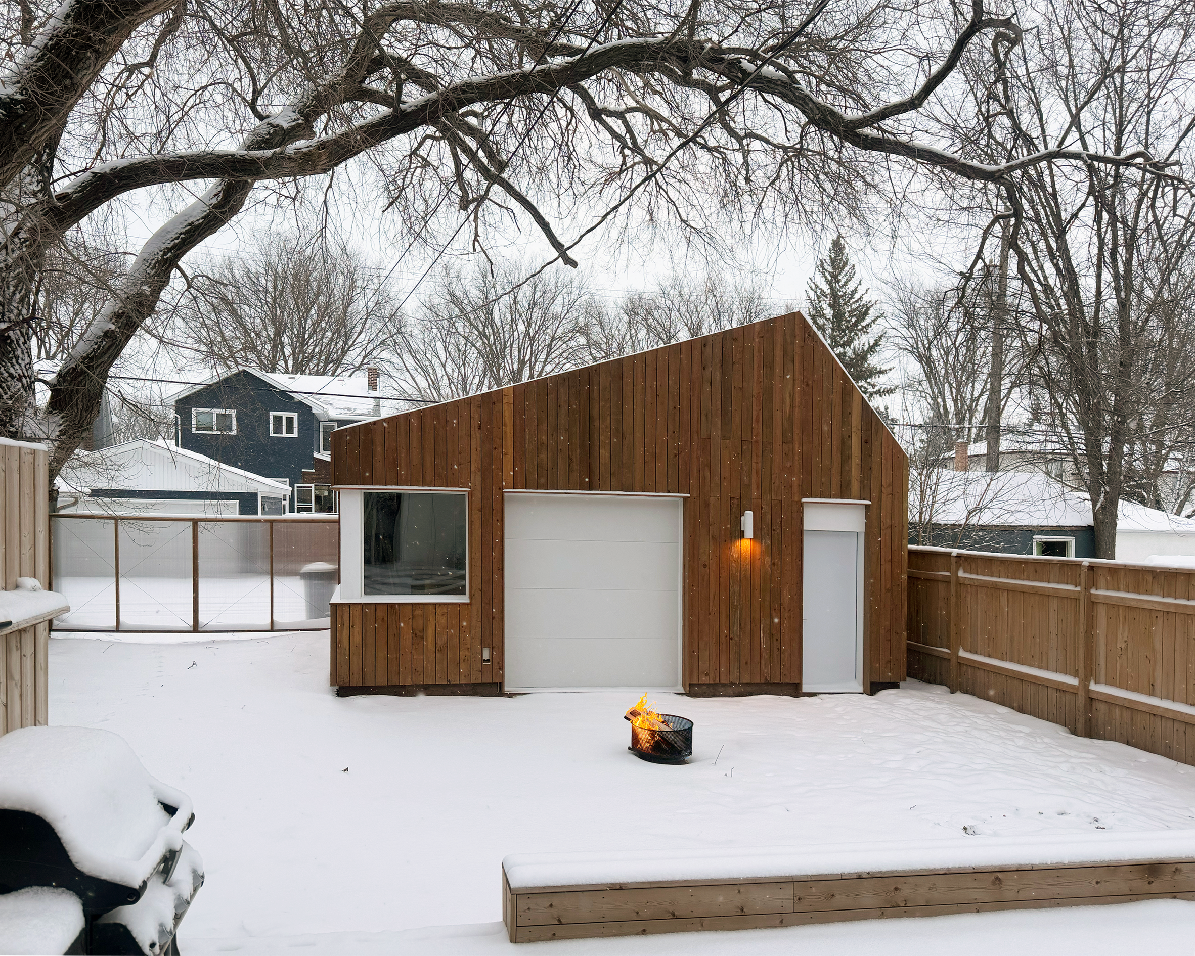 Snow-covered backyard with a small wooden shed, a fire pit with flames, and bare trees in winter. Houses are visible in the background.