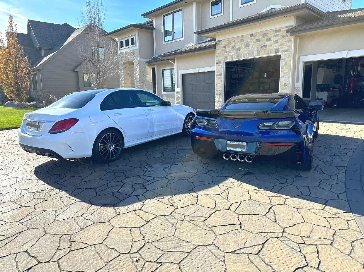 A white luxury sedan and a blue sports car are parked side by side on a stone driveway in front of a modern house with a garage.