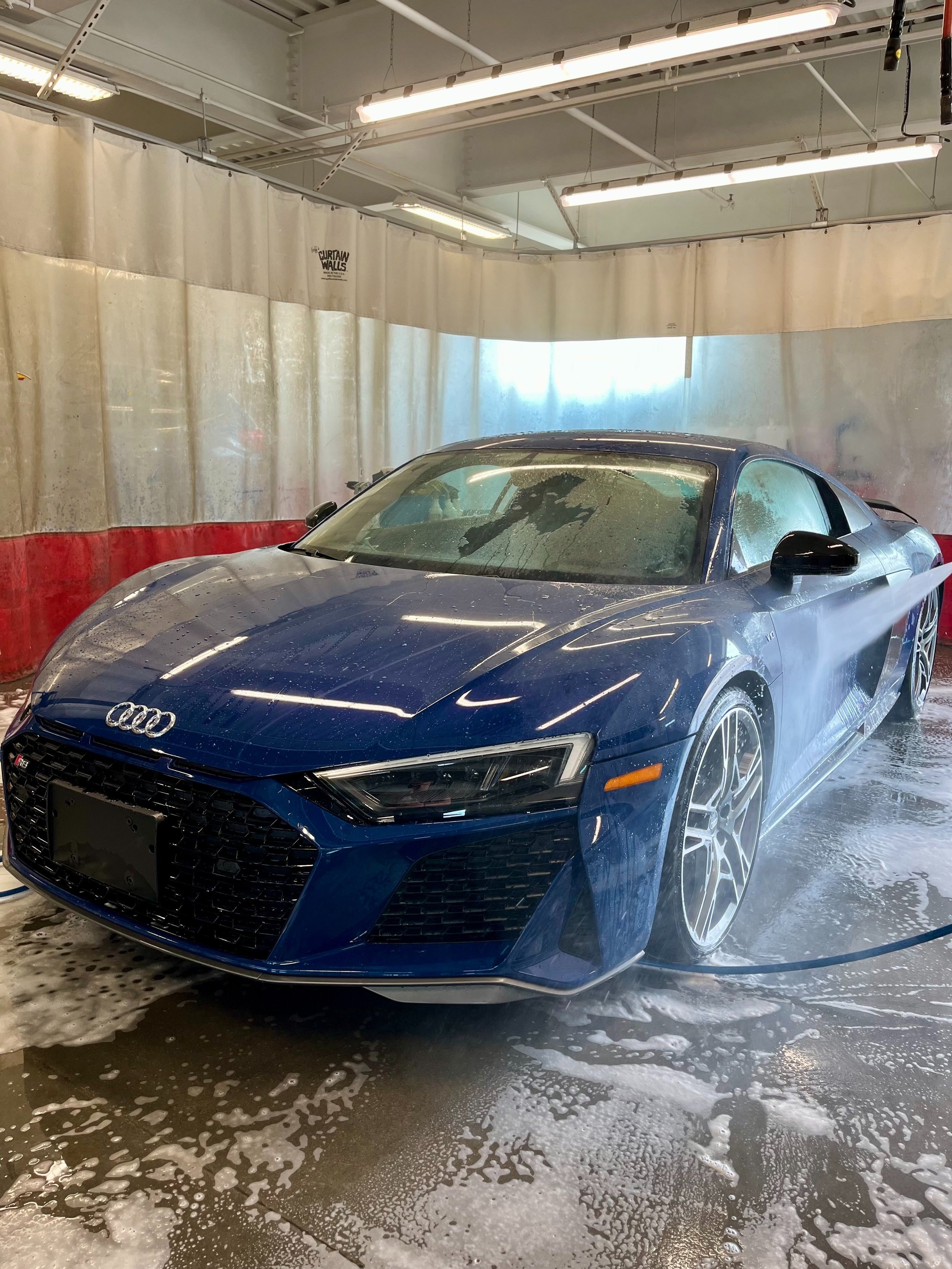 Blue Audi sports car being washed at an indoor car wash station with soap and water spray.