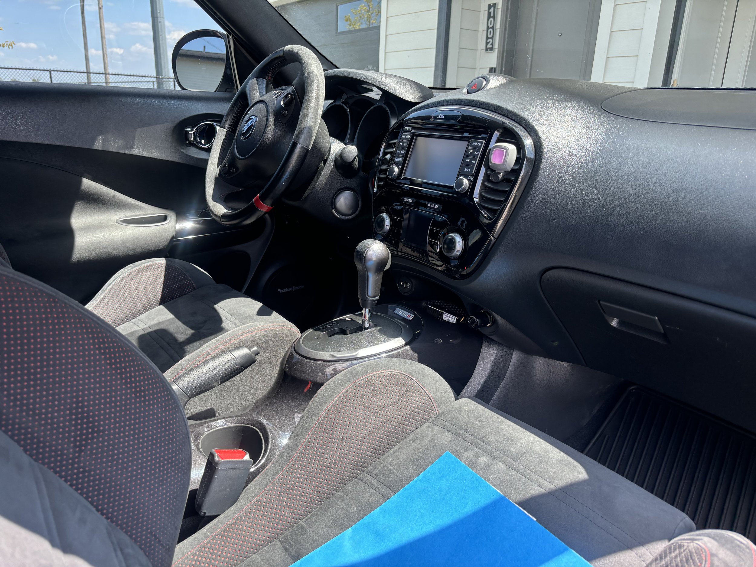 Interior of a compact car showing the dashboard, steering wheel, gear shift, and front seats, with a plastic blue folder on the passenger seat.