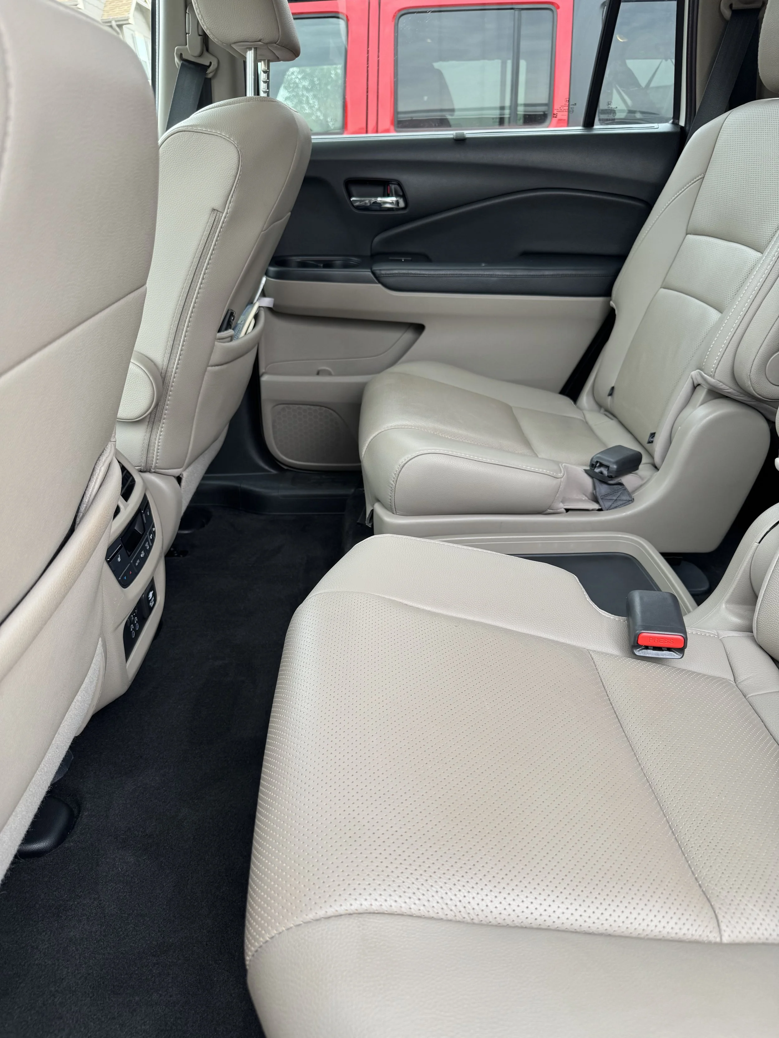 Interior view of a vehicle showing beige leather seats, rear passenger area, with controls and vents at the back of the front seats and a window with a red vehicle visible outside.