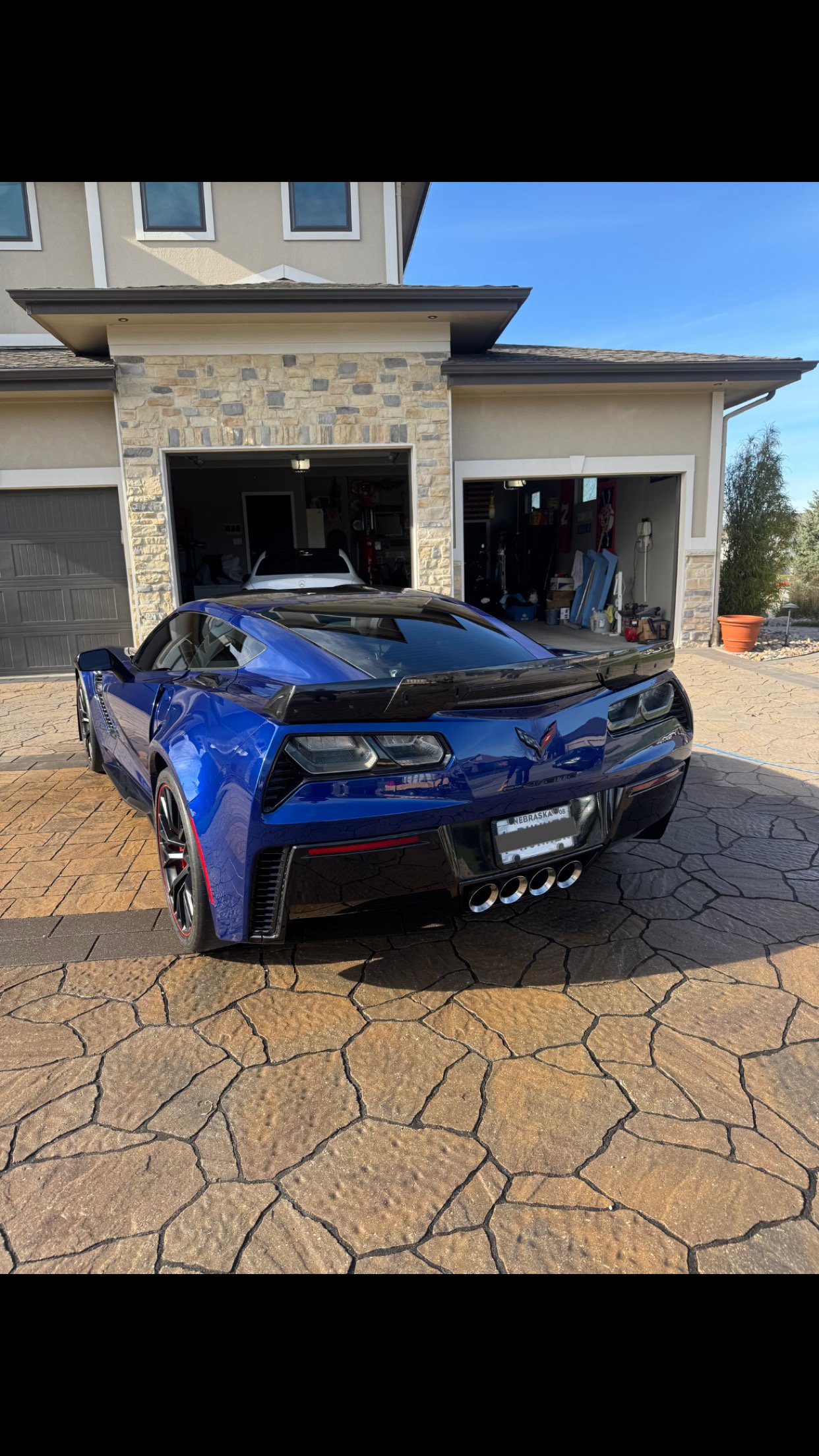 Blue Chevrolet Corvette sports car parked in front of a house with a garage, with other vehicles inside the garage and a stone wall exterior.