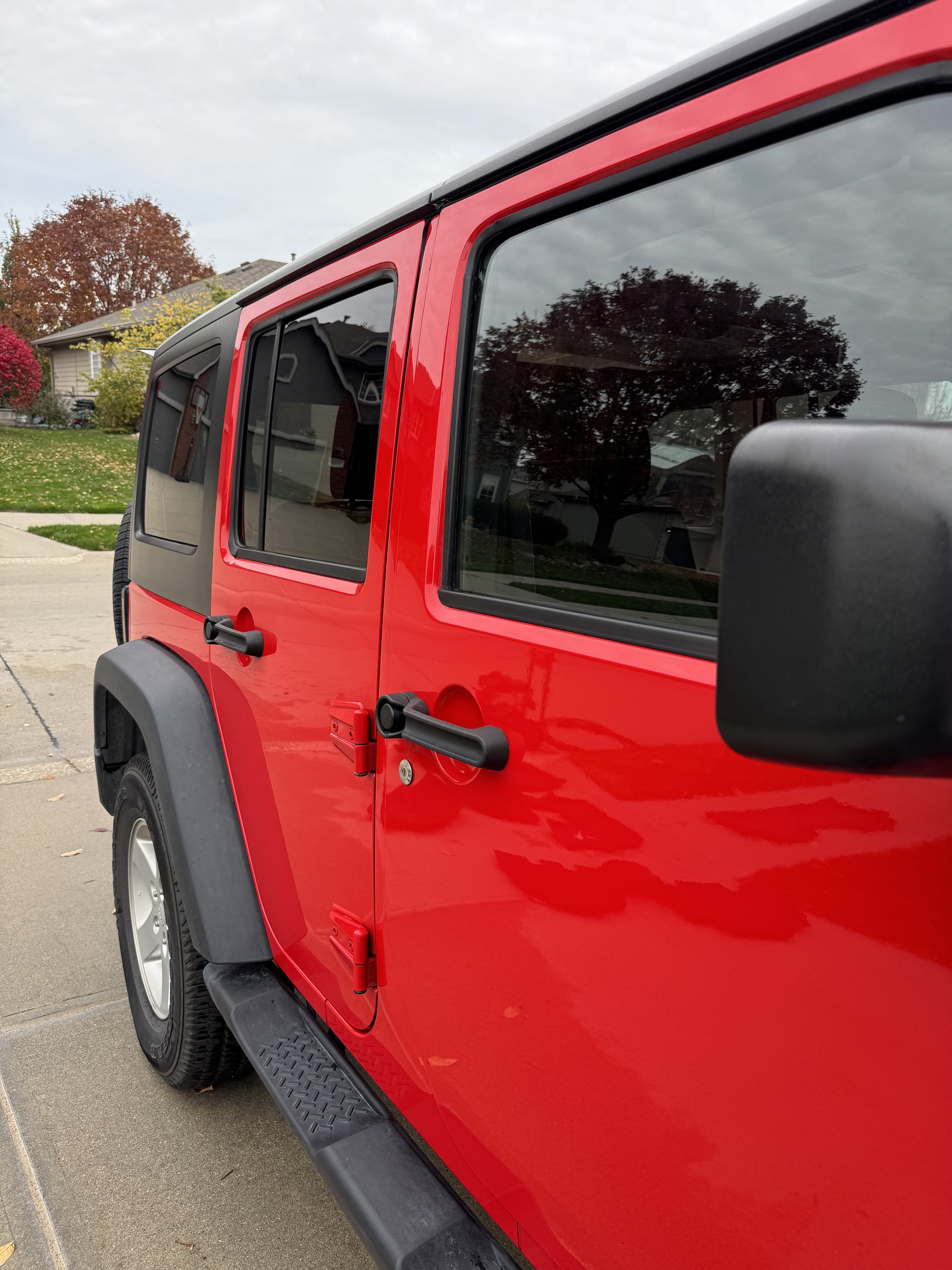 Close-up of red off-road vehicle door with black handles and side mirror, parked on a sidewalk in a suburban neighborhood.