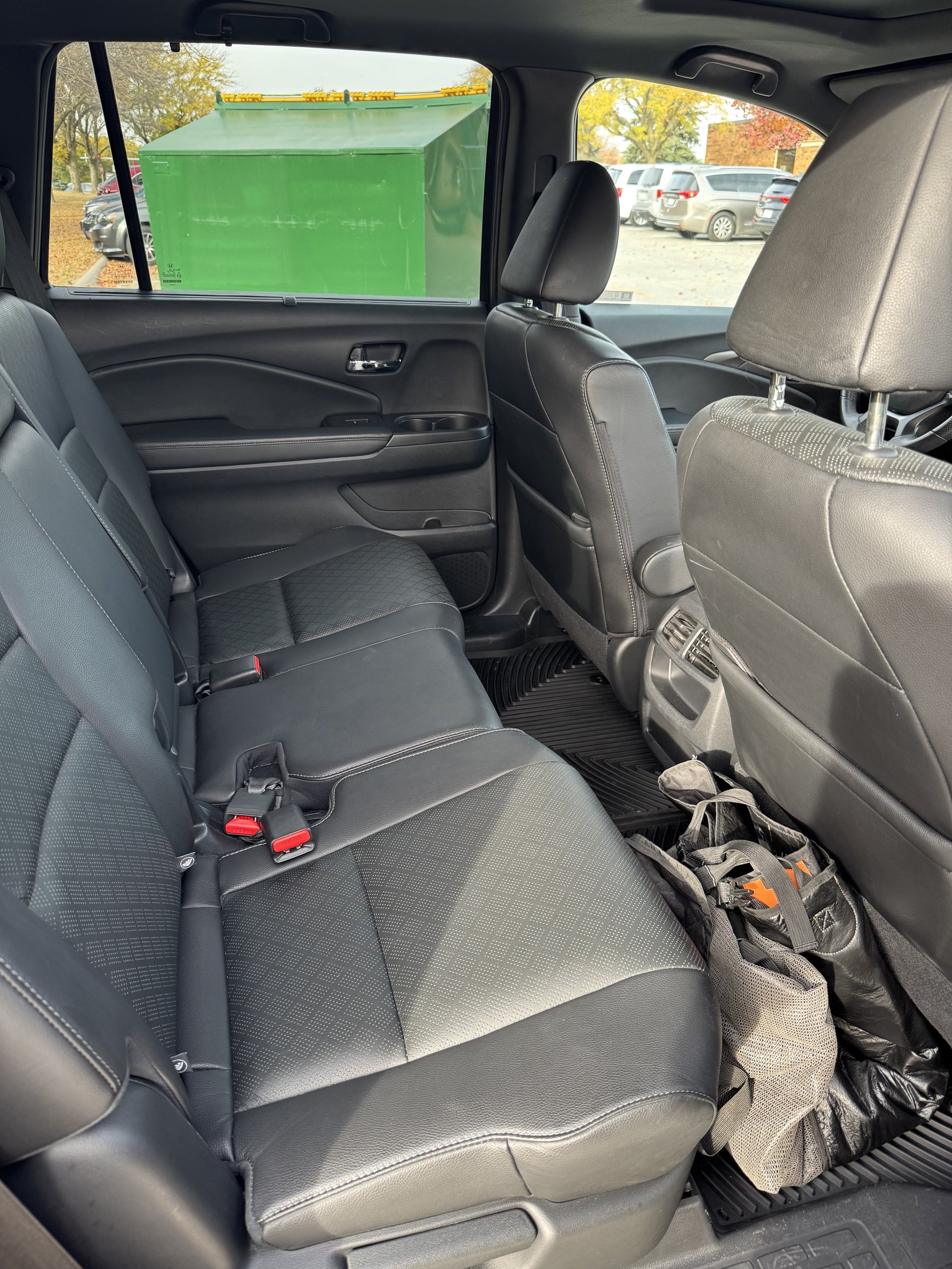 The interior of a vehicle showing the back seat, front seats, and a view outside through the rear window. The back seat has black leather upholstery, and the front seats are also black leather. There is a green container visible through the rear wind
