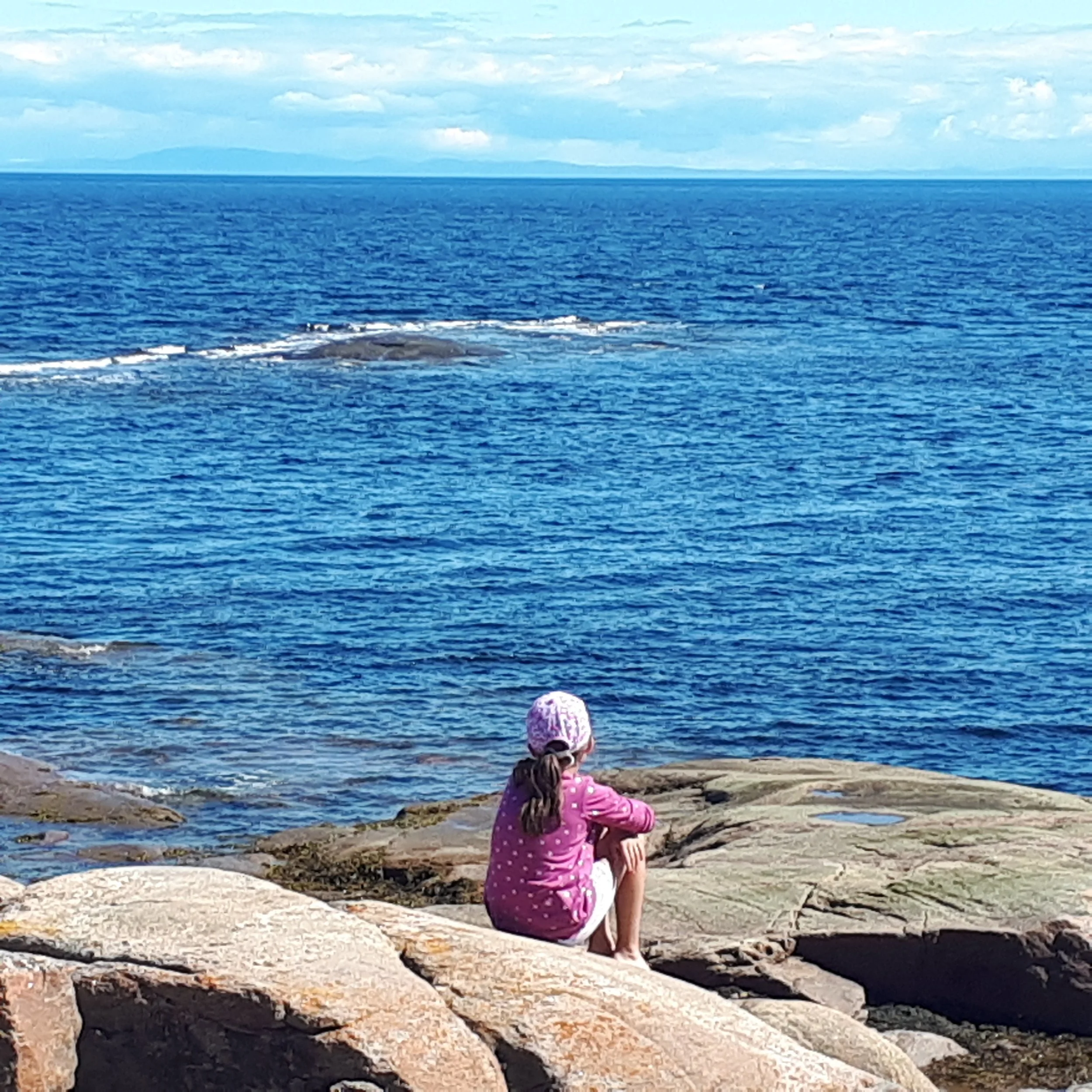 Une jeune fille assise sur des rochers regardant la mer, portant un chapeau rose et un manteau rose avec des pois blancs.