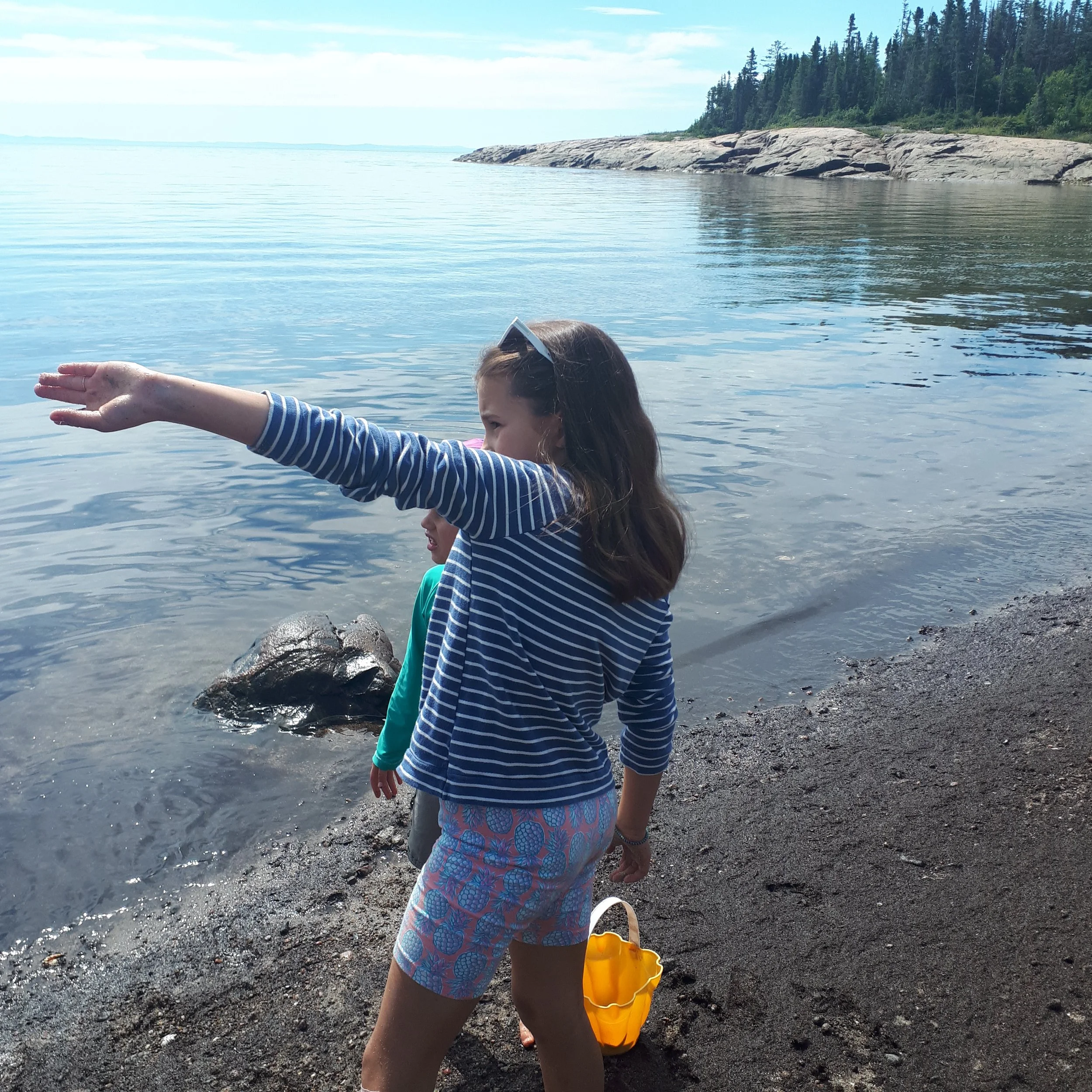 Une fille en t-shirt à rayures bleues et blanches, shorts à motifs ananas, pointe vers le lac, avec un seau jaune à ses pieds, plage rocheuse et forêt en arrière-plan.
