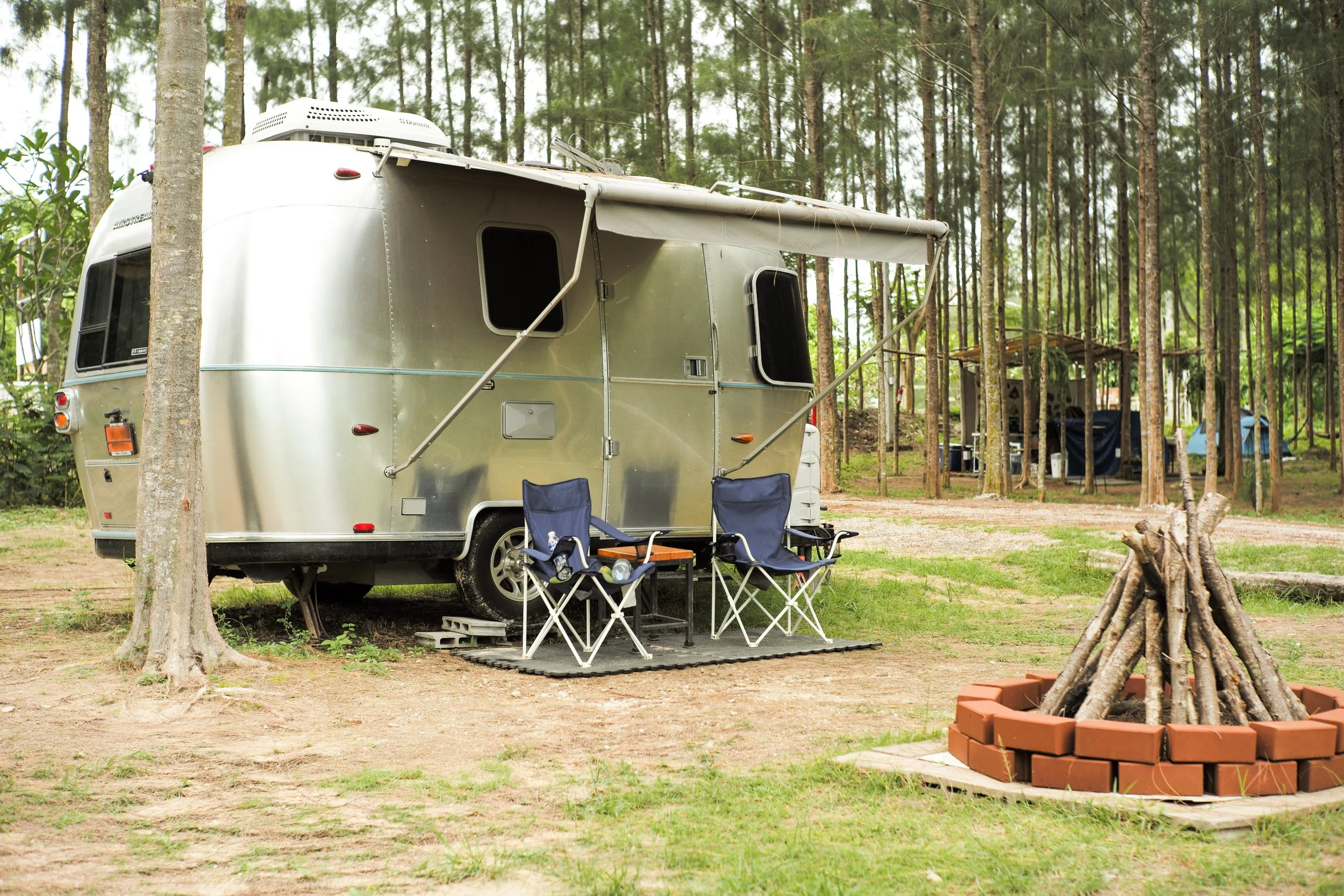 Caravane en camping entourée d'arbres, avec deux chaises pliantes, une petite table, et un feu de camp avec des bûches en briques.