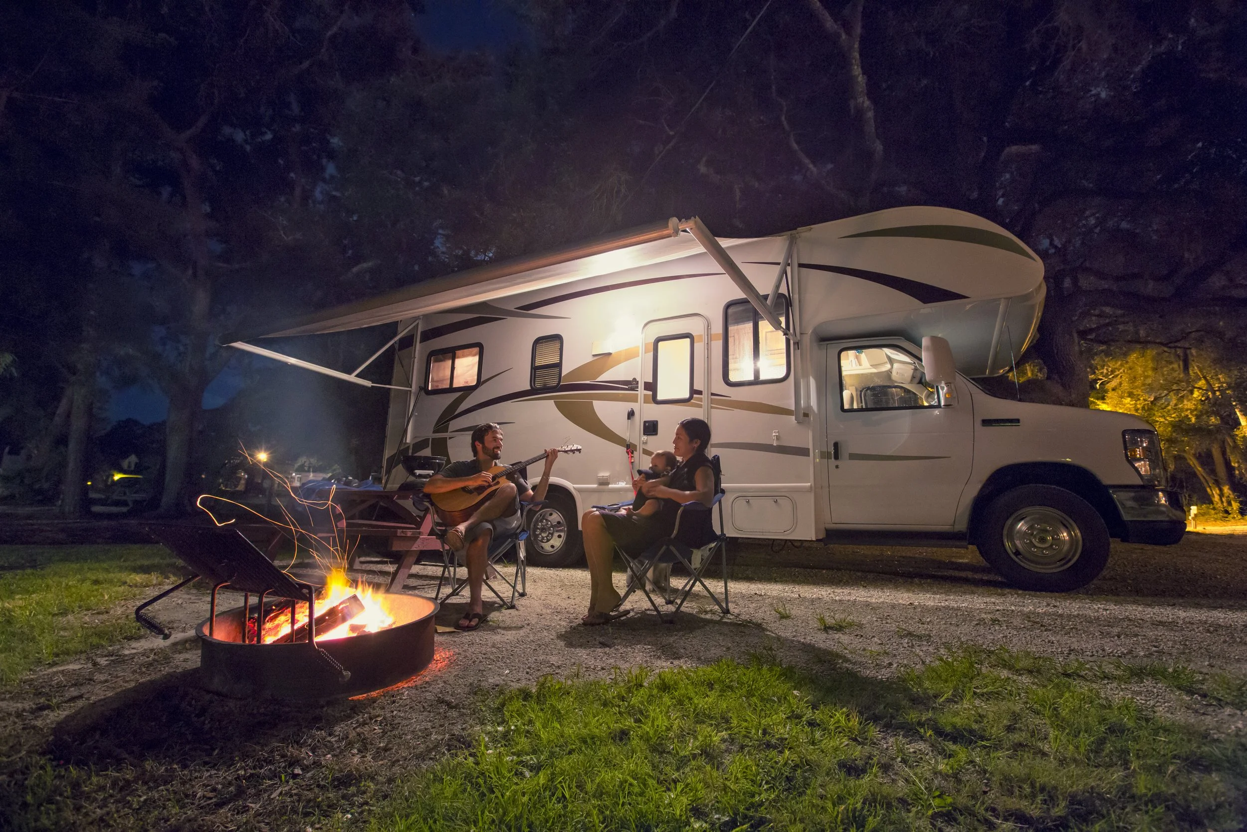 Famille profitant d'une soirée autour d'un feu de camp à côté d'une caravane dans un camping en forêt nocturne.