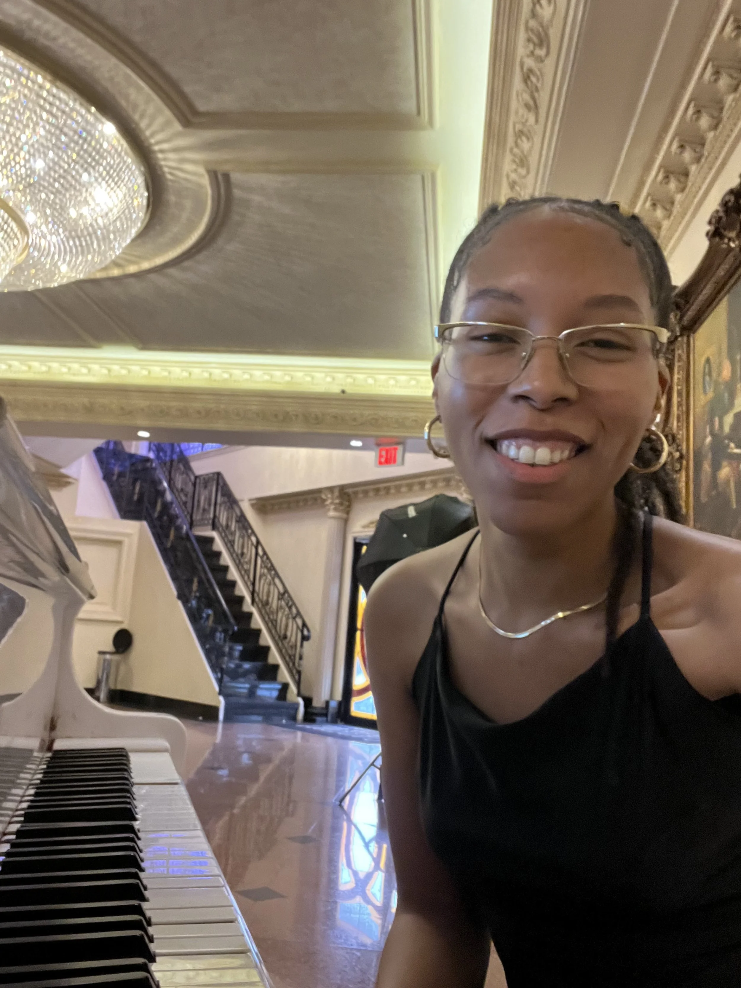 Smiling person in a formal setting with a piano and chandelier