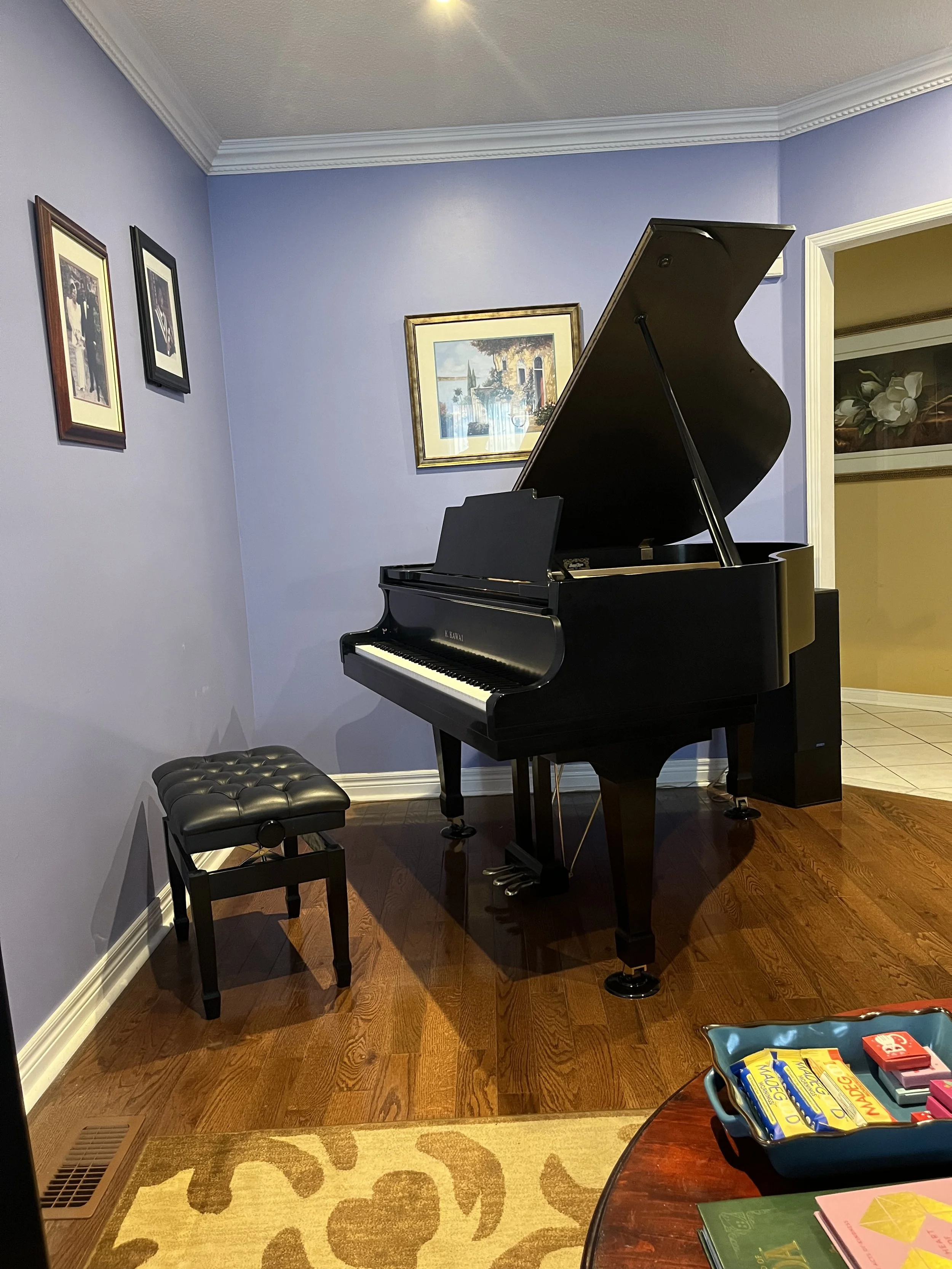 Grand piano in a room with purple walls, framed pictures on the wall, a black piano bench, and various items on a nearby table.