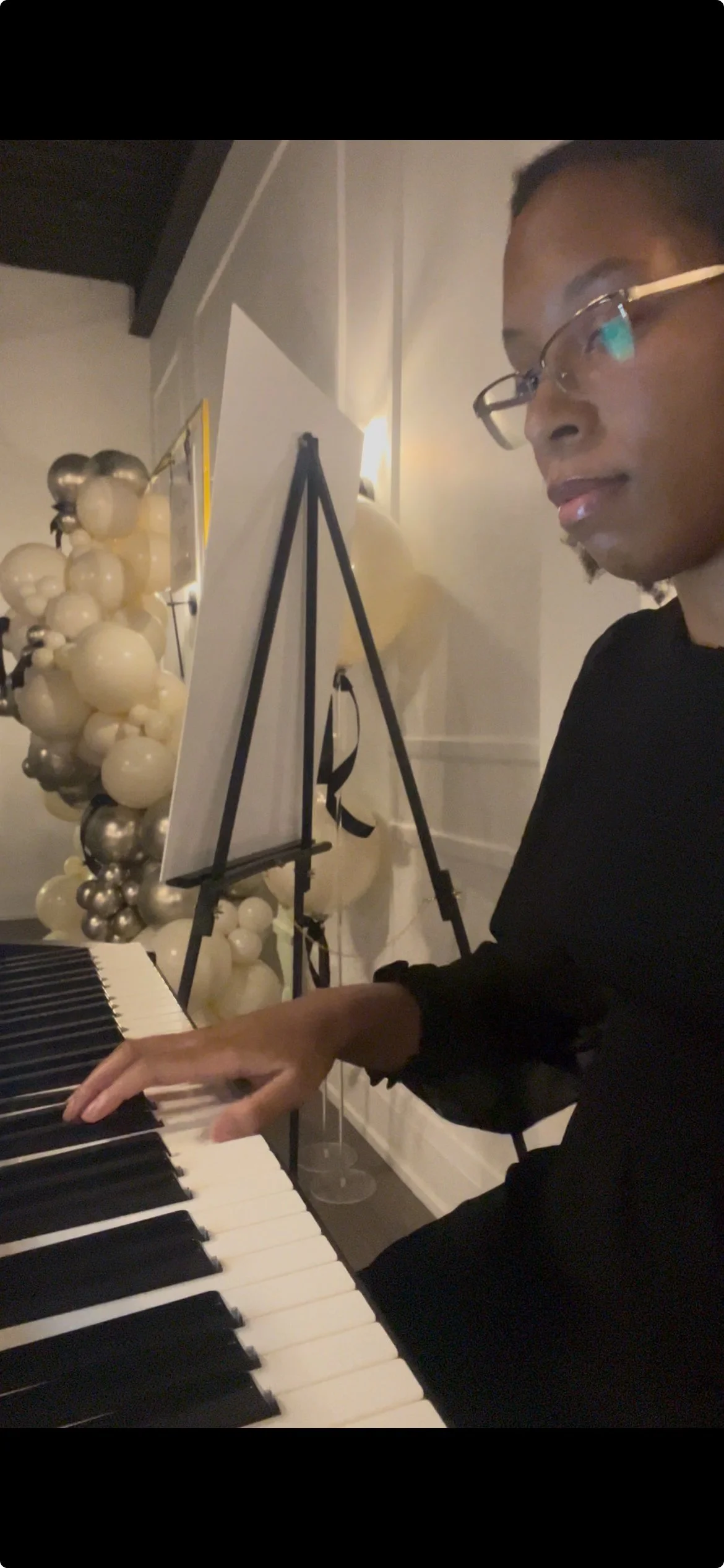 Person playing a keyboard at an event with a balloon decoration and easel in the background.