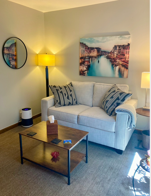 Living room with beige sofa decorated with two striped pillows, a wooden coffee table with a tissue box and a small decorative item, a standing lamp, a wall art of a canal scene, and a round mirror on the wall.