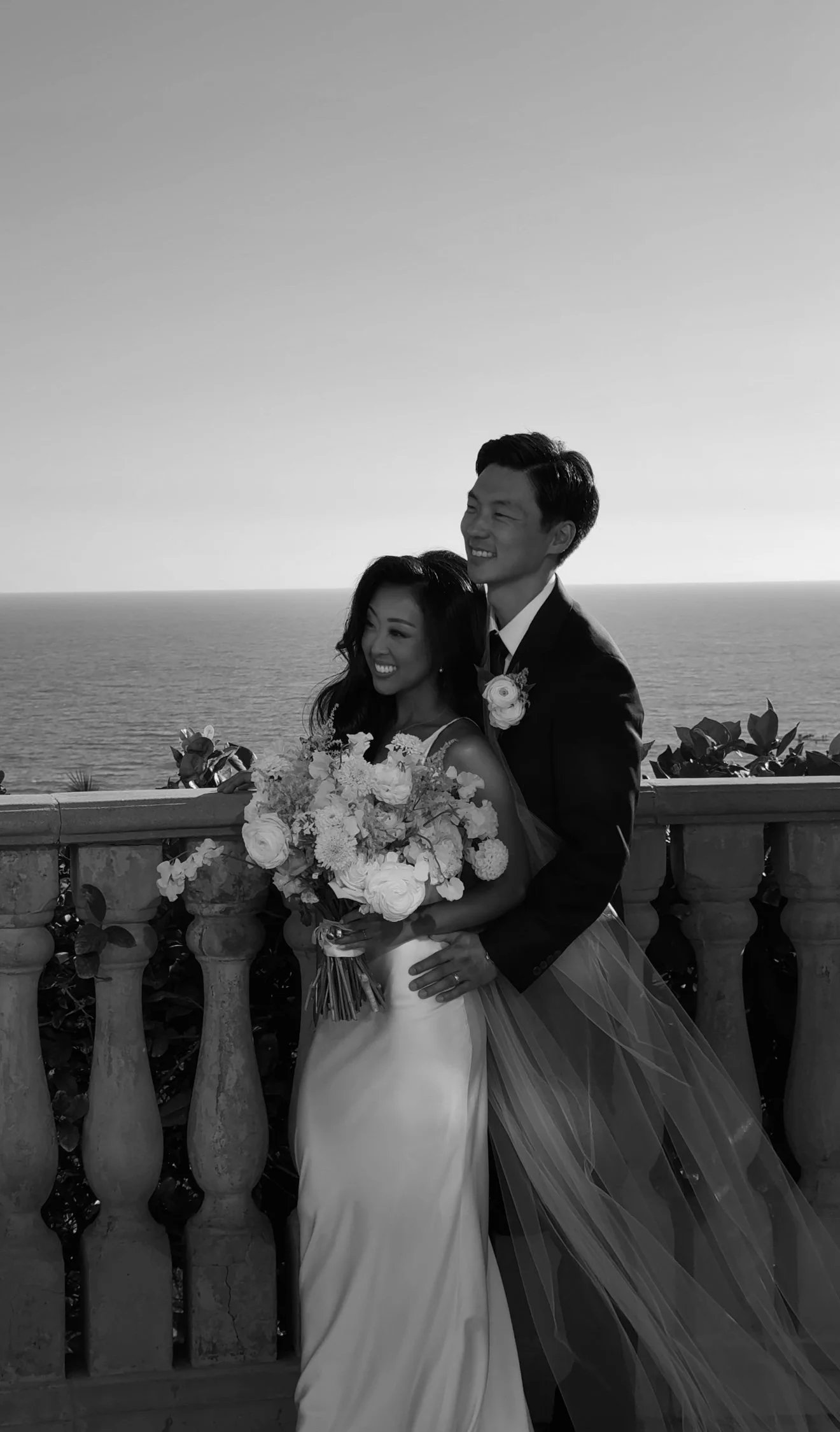 Black and white photo of a couple walking by a reflective pool, dressed in formal wedding attire; the groom in a tuxedo and the bride in an off-shoulder gown, with a forested background.