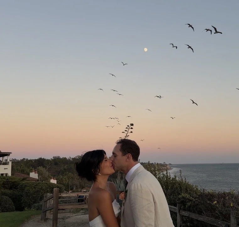 A couple sharing a kiss outdoors at sunset with the ocean in the background, flying birds, and a full moon in the sky.