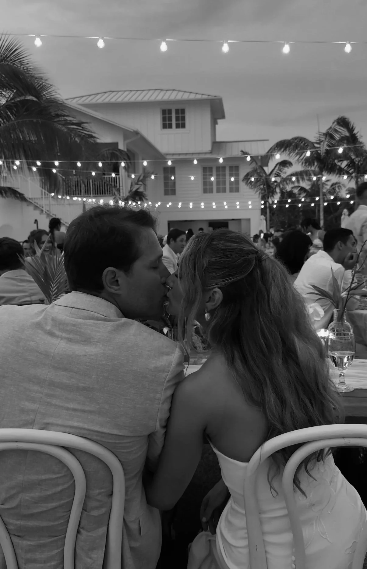 A black and white photo of a couple kissing at an outdoor event with string lights, palm trees, and a house in the background.