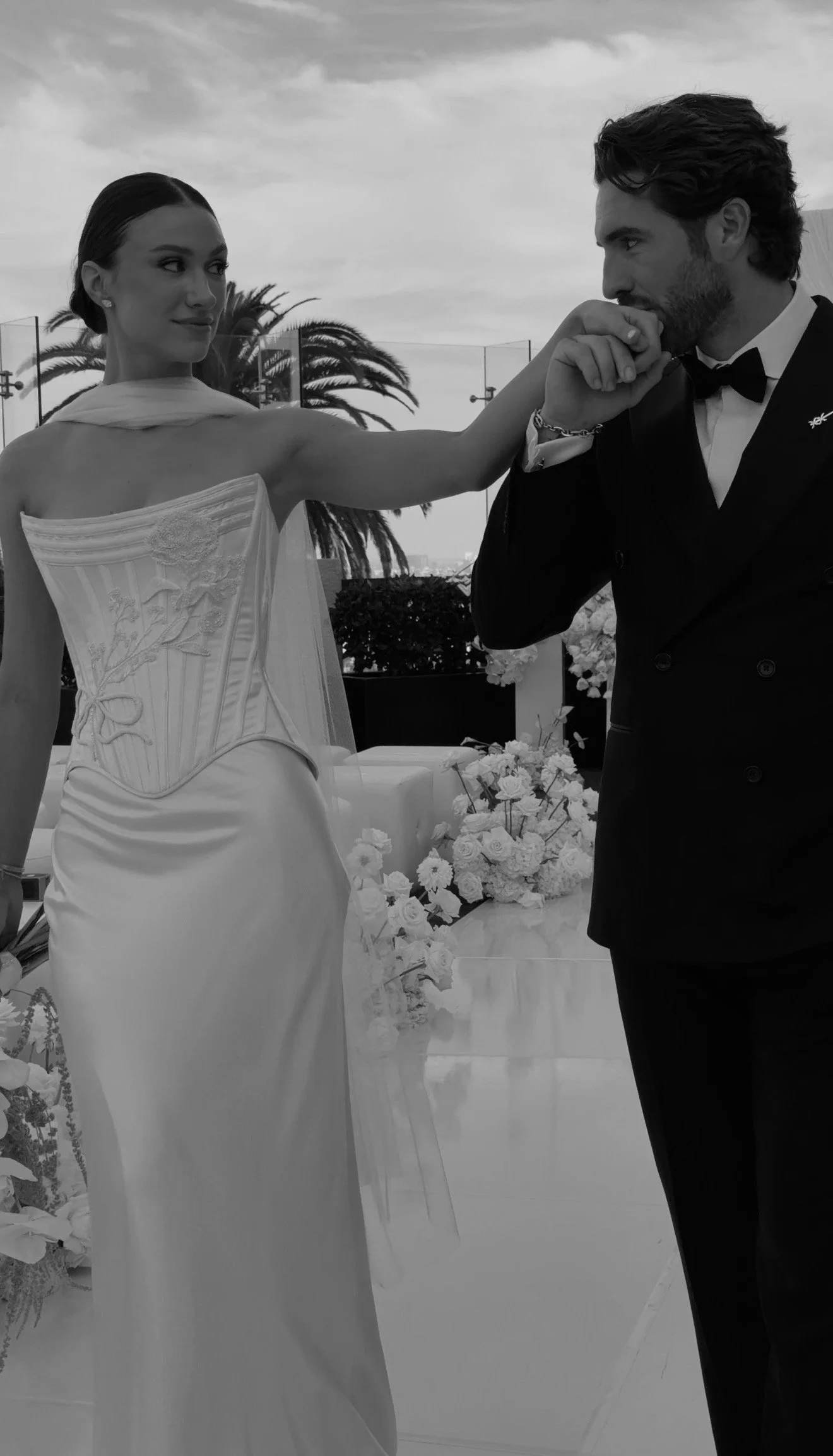 Bride with veil holding white flowers surrounded by blurred guests on a checkered floor at a wedding.