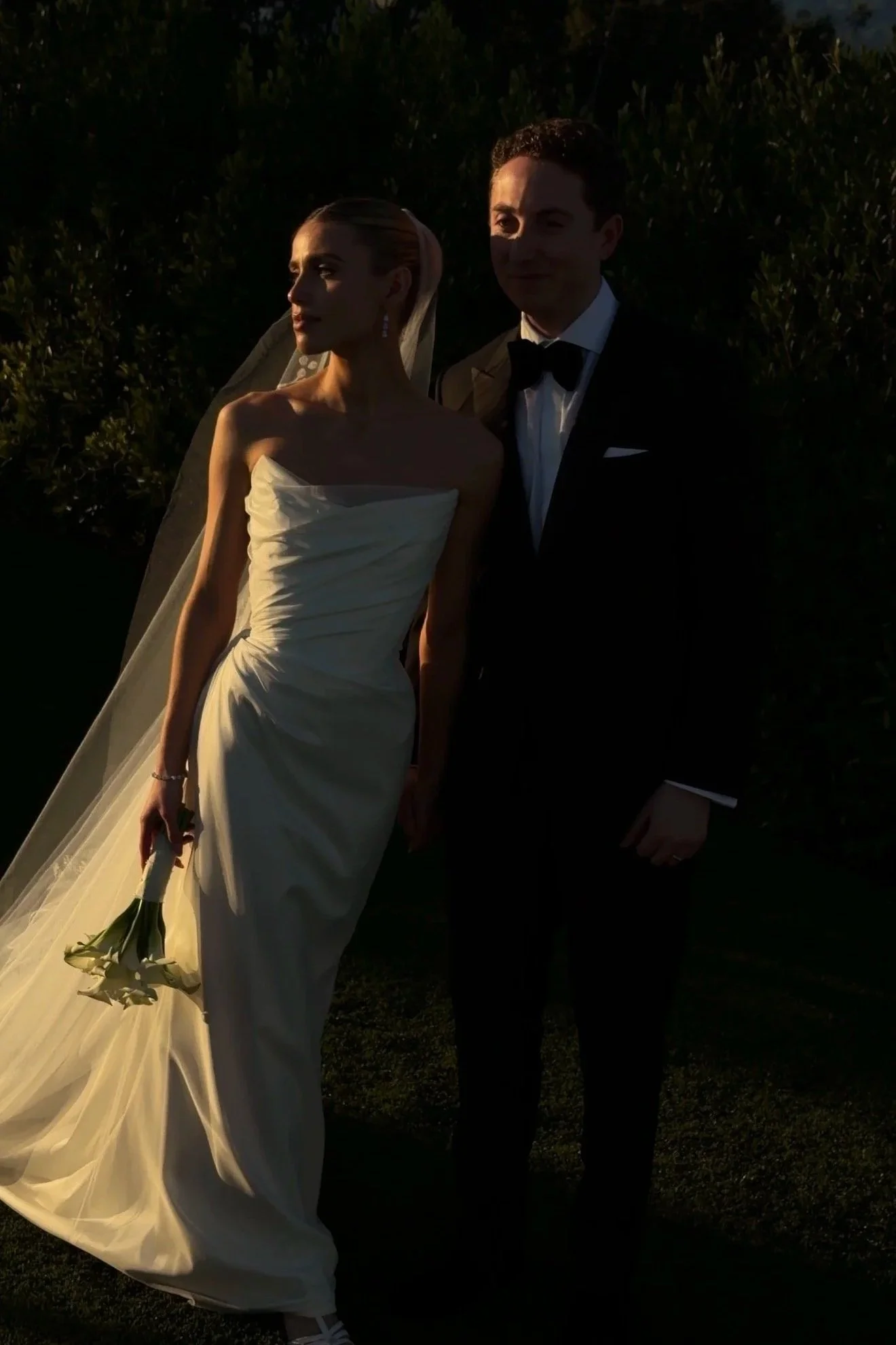 A bride and groom dressed in wedding attire, standing outdoors during sunset. The bride holds a bouquet of white calla lilies and has her hair styled elegantly. The groom is wearing a black tuxedo with a bow tie. Both are partly illuminated by the setting sun.