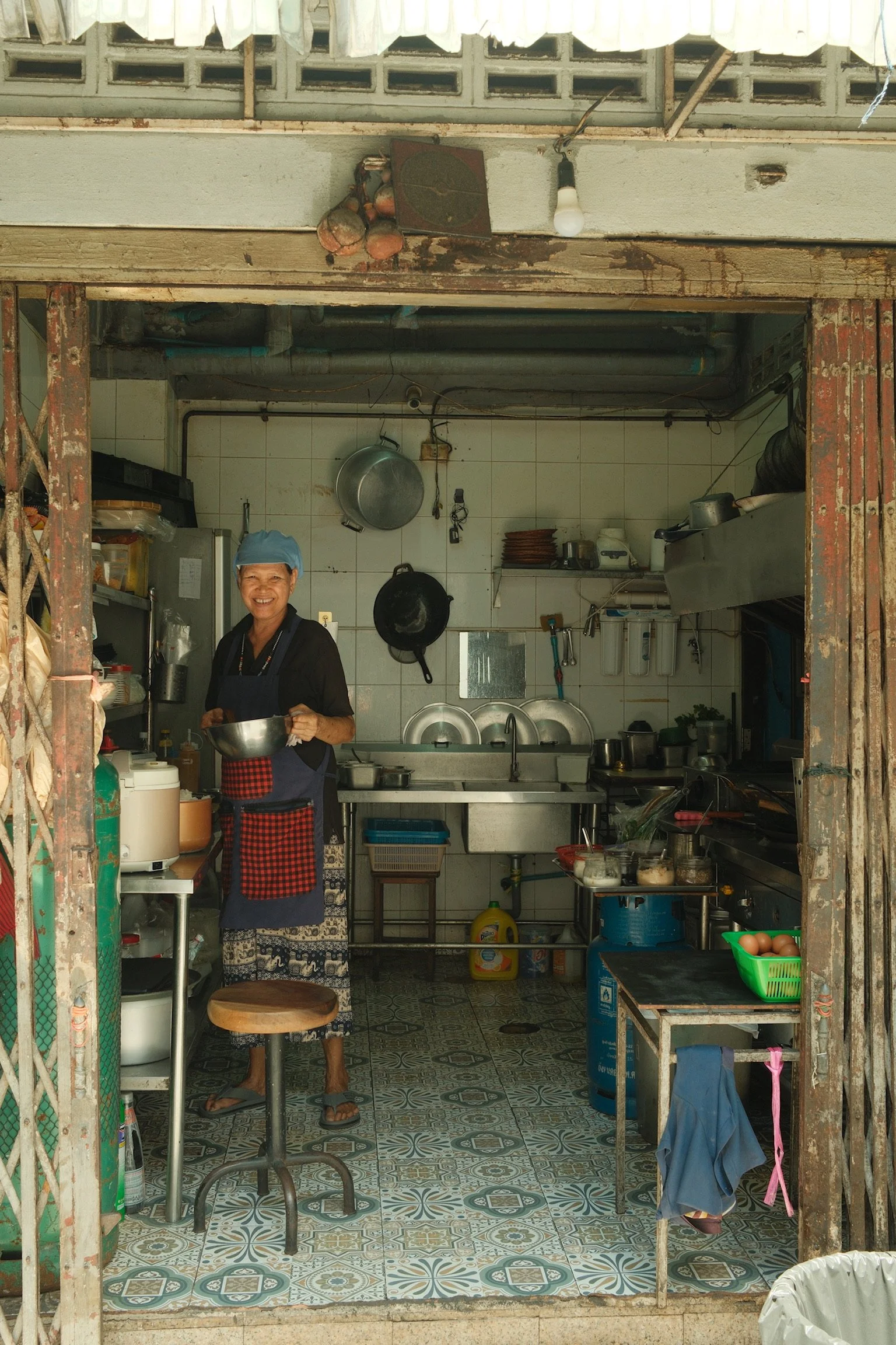 Een vrouw staat in een kleine, eenvoudige keuken en lacht naar de camera. Ze houdt een metalen kom vast. De keuken heeft witte tegels, hangende pannen, en diverse keukengerei en ingrediënten op het aanrecht.