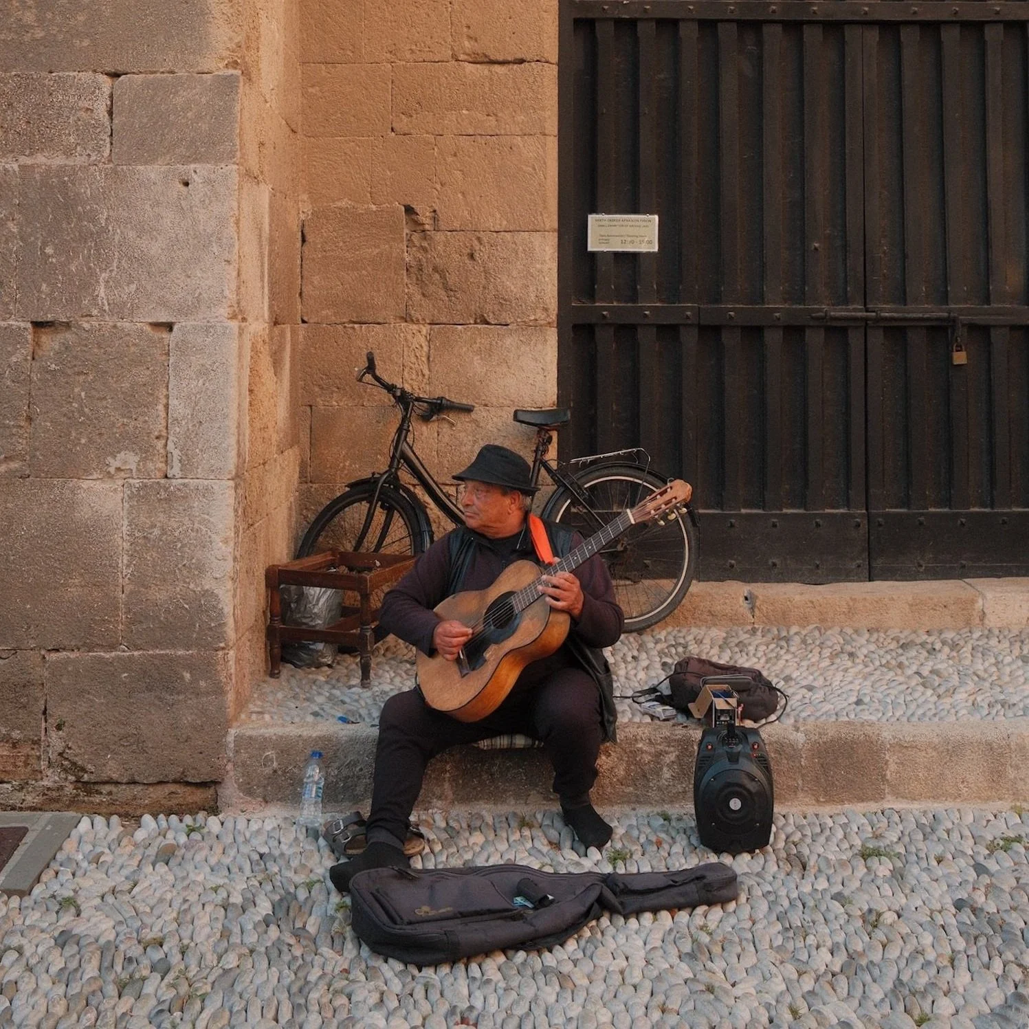 Straatmuzikant speelt gitaar voor een historische stenen muur, met een fiets op de achtergrond en zijn spullen op straat.