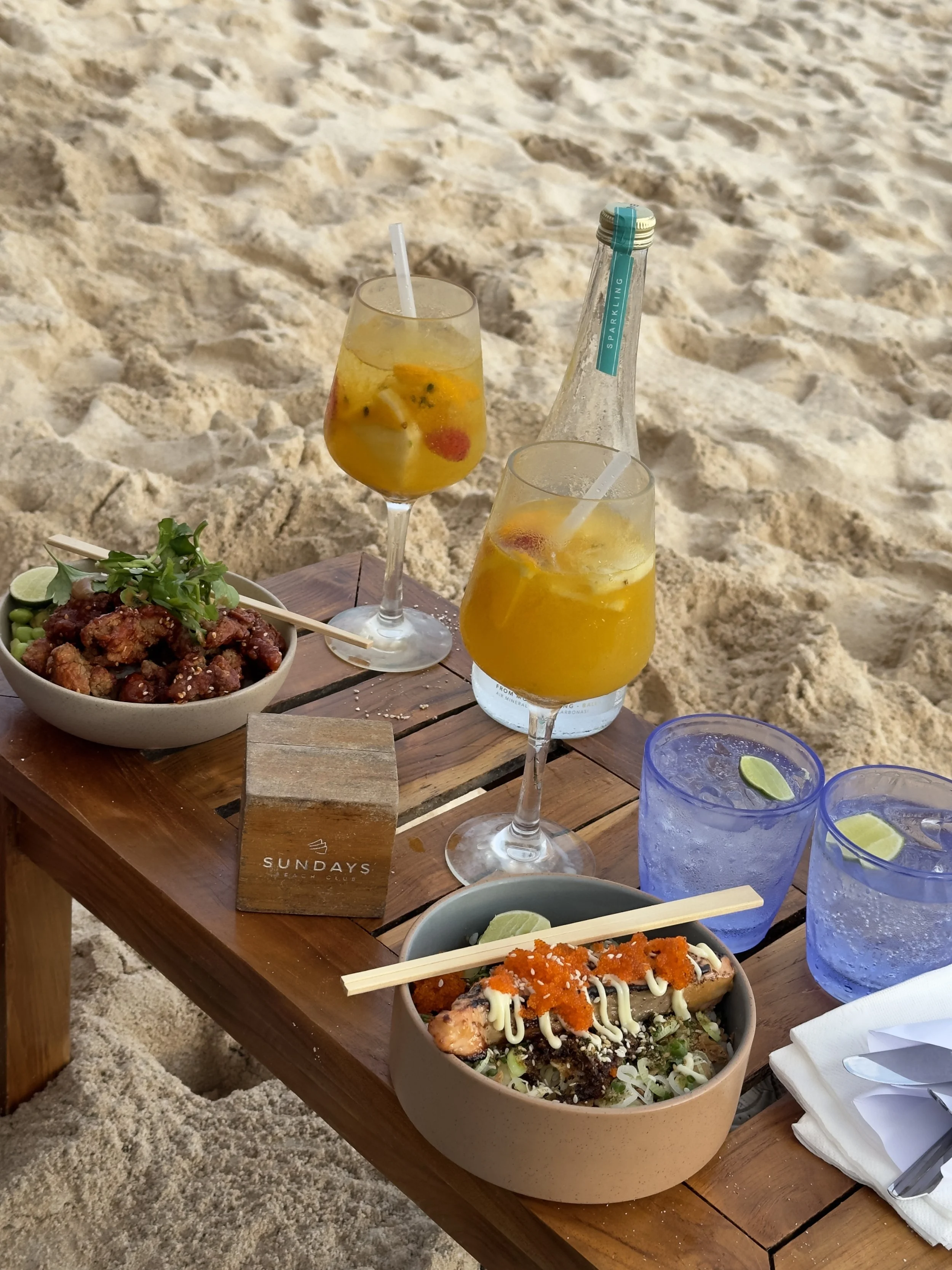 Een onderzetter met het logo 'SUNDAYS BEACH CLUB' op een houten tafel met sushi, fruitige cocktails, en drankjes op het zand.