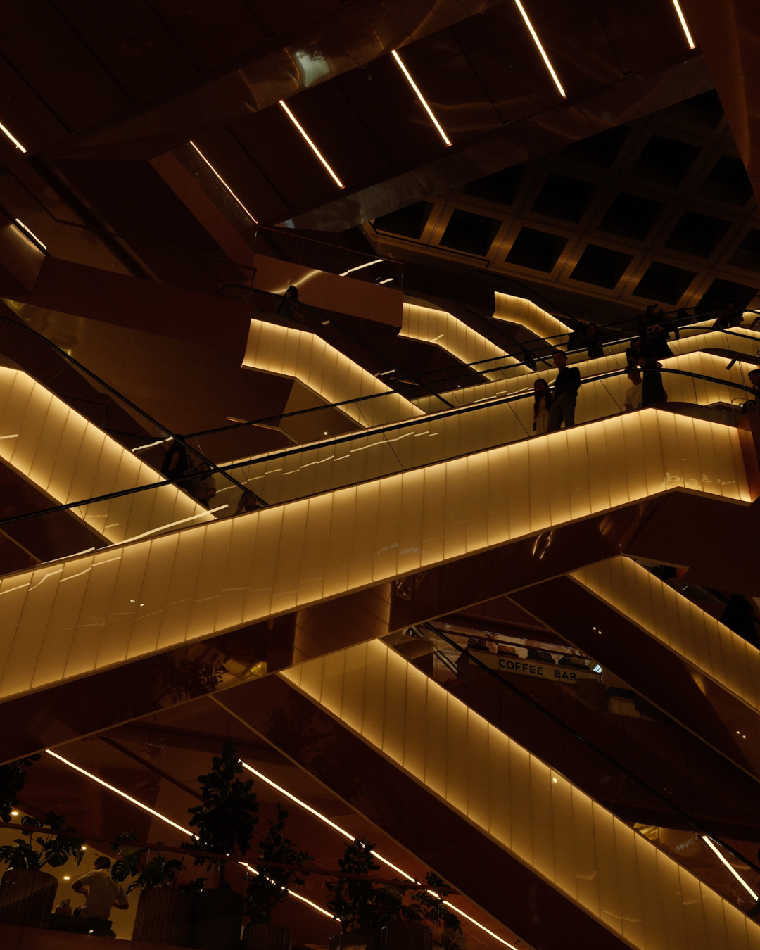 Interieur van een modern winkelcentrum  met meerdere verdiepingen en escalators, verlicht met warme gele verlichting, mensen lopen op de escalators.