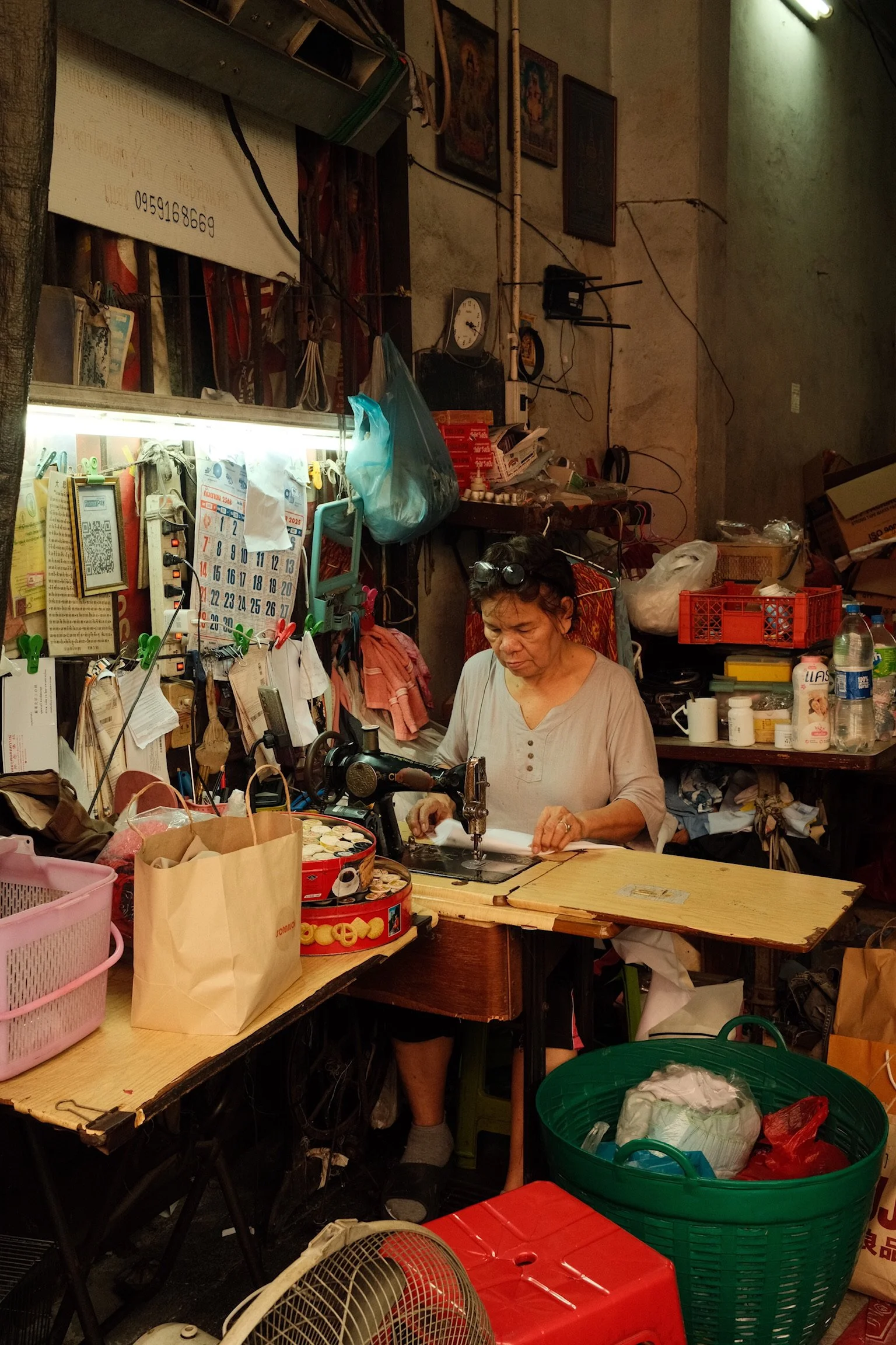 Een vrouw naait kleding of stof in een drukke werkruimte vol spullen, zoals dozen, tassen, en werkmateriaal.