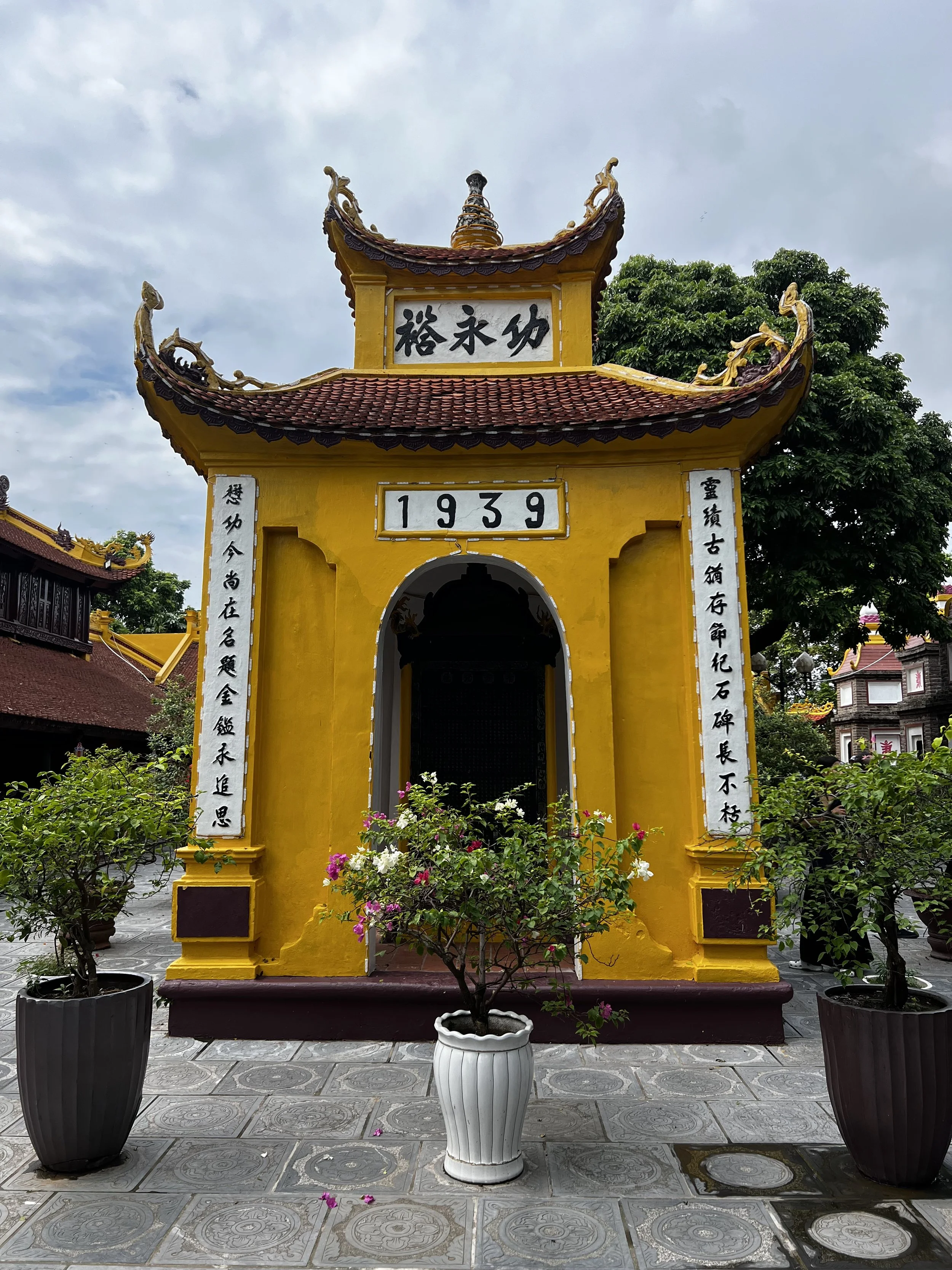 Een geel pagodetekening uit 1939 met rode, gekrulde dakranden en Chinese karakters, omgeven door planten en potten.