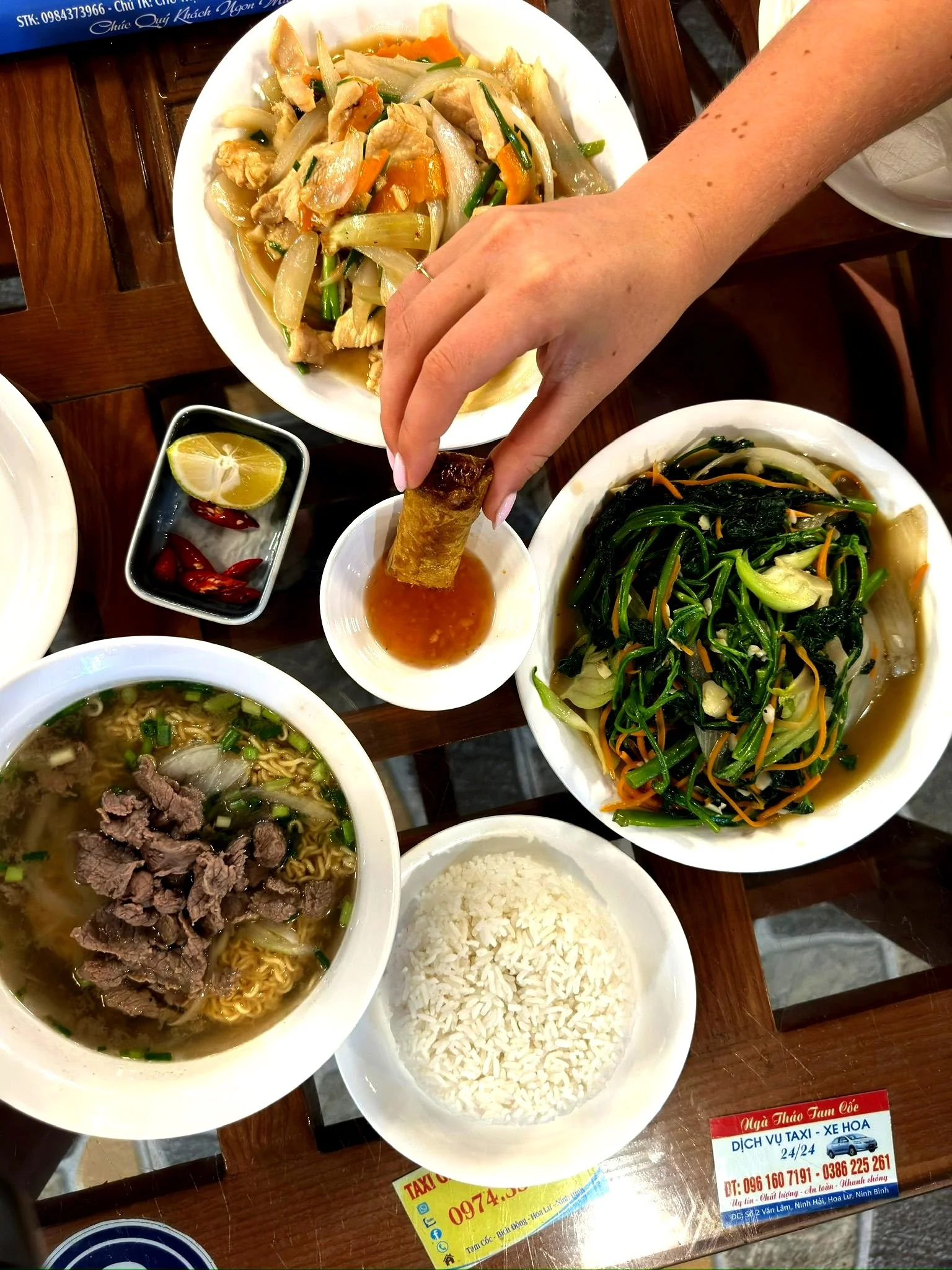 Diverse Vietnamese gerechten op een tafel, inclusief rijst, noedelsoep met rundvlees, roergebakken groenten en een plateet met een vleesgerecht. Er wordt een lenterol in een saus gedipt.