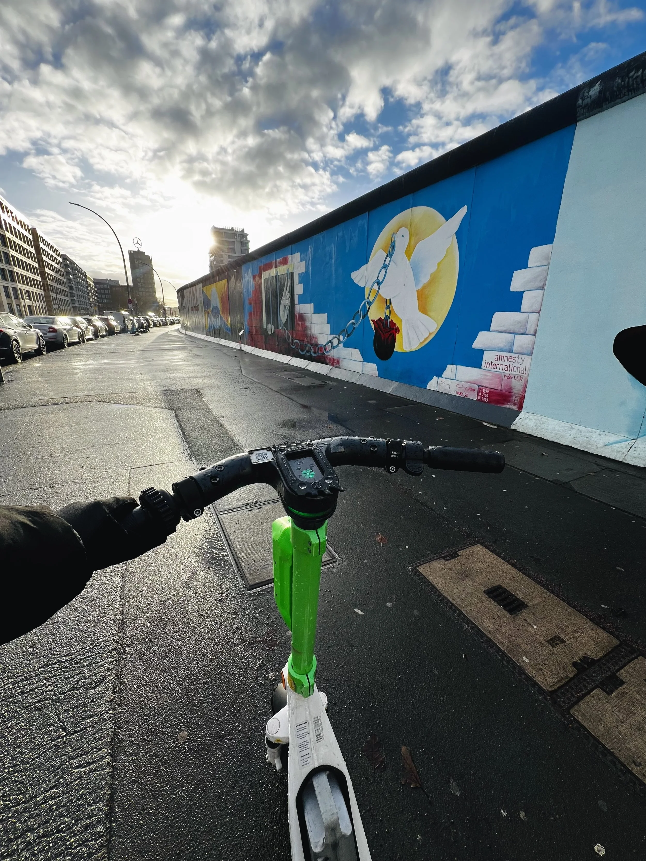 Uitzicht vanaf een scooter op een straat met een muur en muurschildering met een duif en een geabstraheerd hart, onder een bewolkte hemel met zon op de achtergrond.