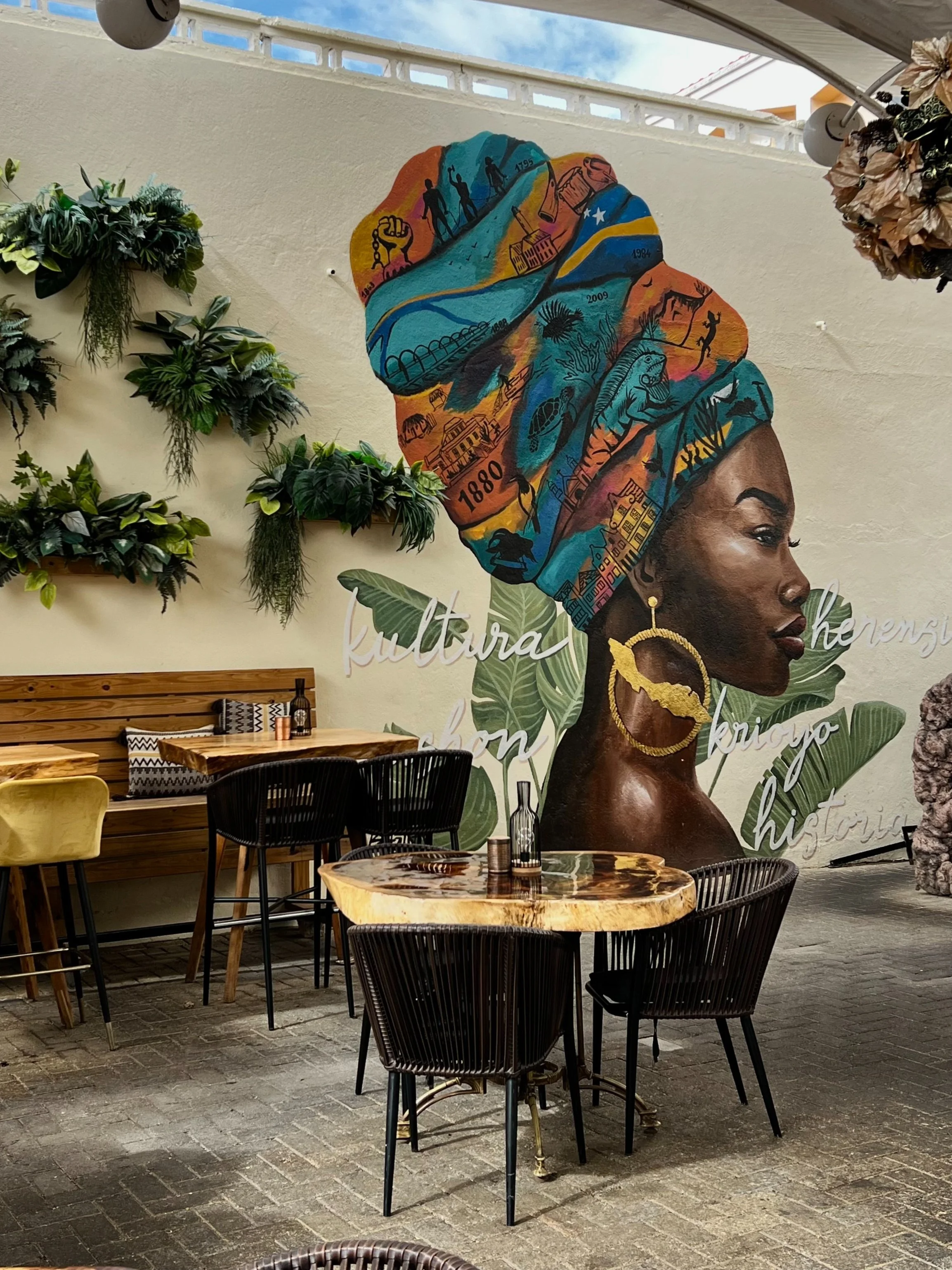 Muur graffiti kunstwerk van een vrouw met een kleurrijke hoofddoek, omringd door groene planten en interieurmeubilair in een restaurant of café.