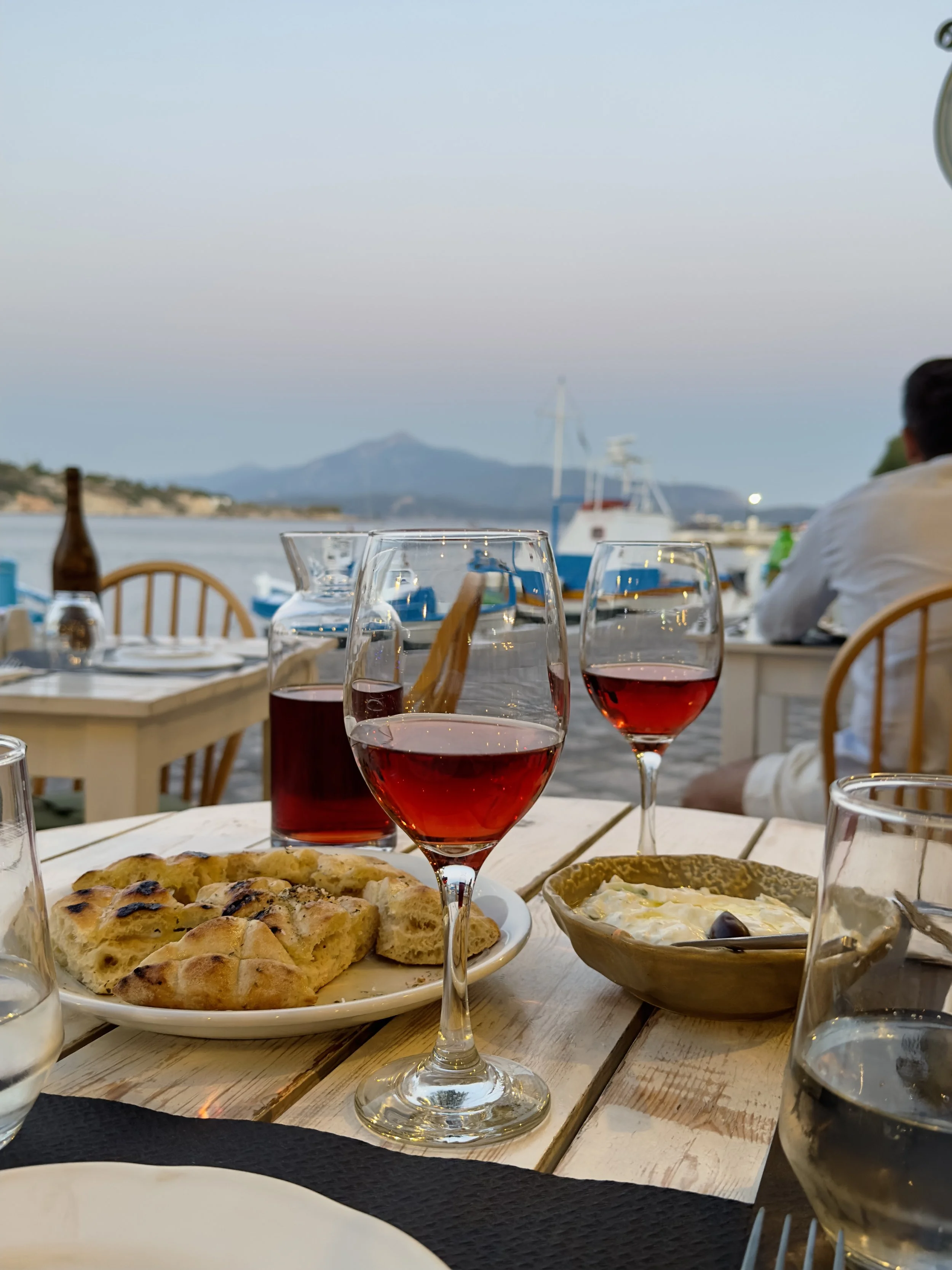 Een tafel met drie glazen rosé wijn, een bord met gegrilde flatbread, een kom met een salade, en een paar glazen water. Op de achtergrond is een zee met boten en een berg in de verte.