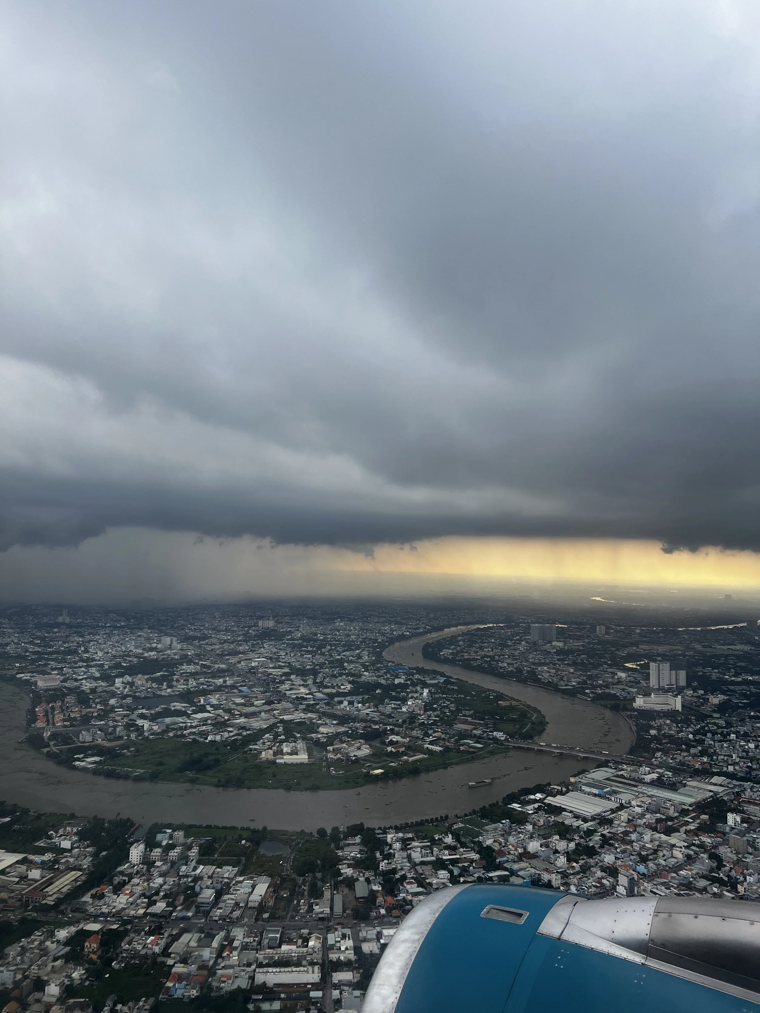 Luchtfoto van een stad onder een bewolkte hemel, met een rivier die door de stad stroomt en een vliegtuigvleugel op de voorgrond.