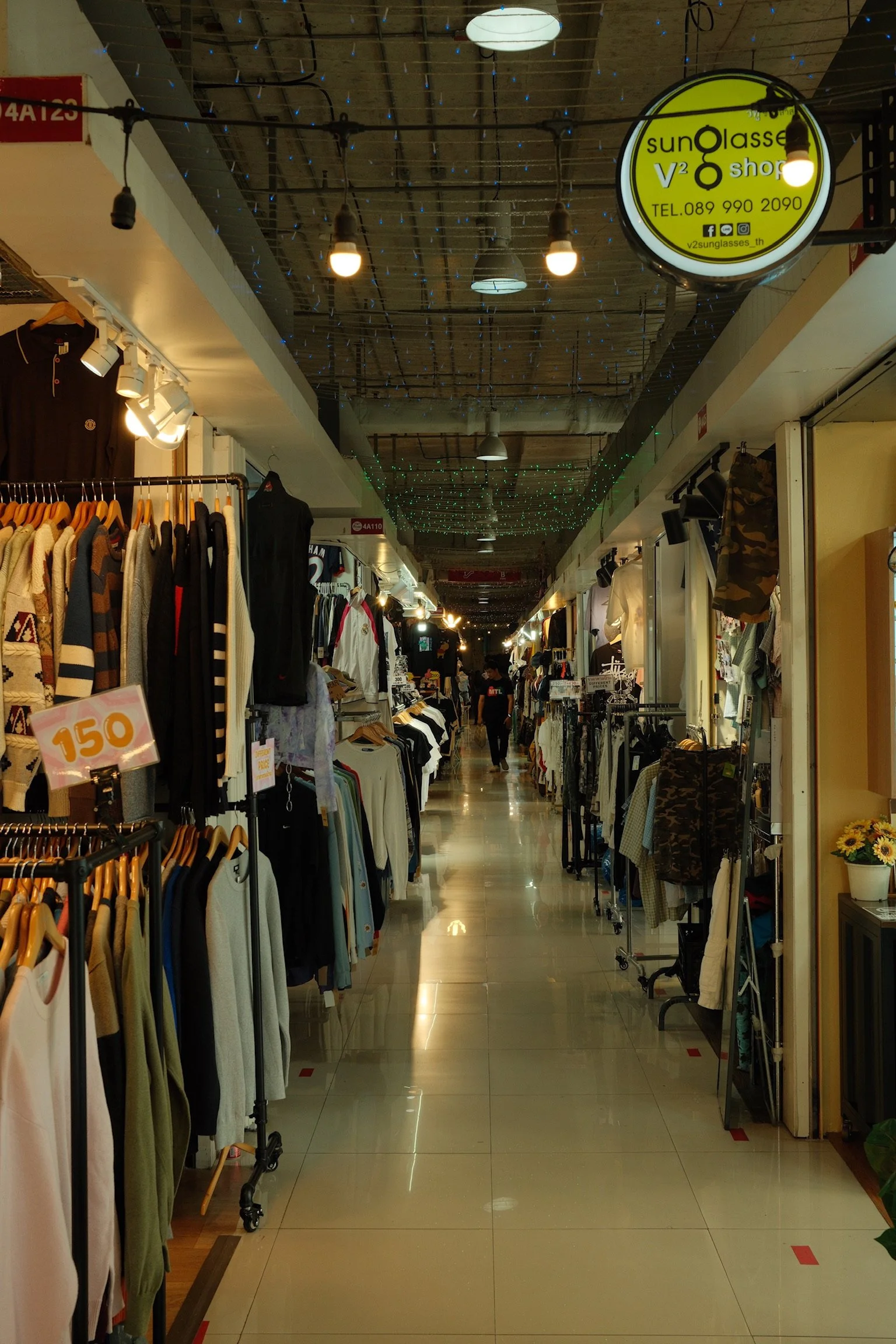 Binnenkant van een winkelpassage met kledingrekken en winkels aan beide zijden, enkele mensen lopen door de gang, verlichte plafond met decoratieve draadjes en hangende lampen.