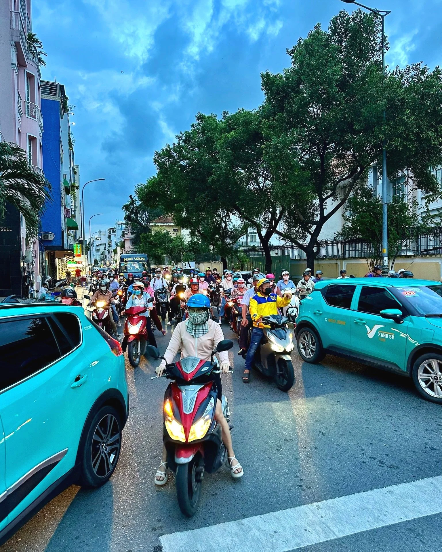 Straat vol met scooters en enkele auto's, met veel mensen die dragen helm en mondkapjes, onder een bewolkte lucht met grote bomen op de achtergrond.