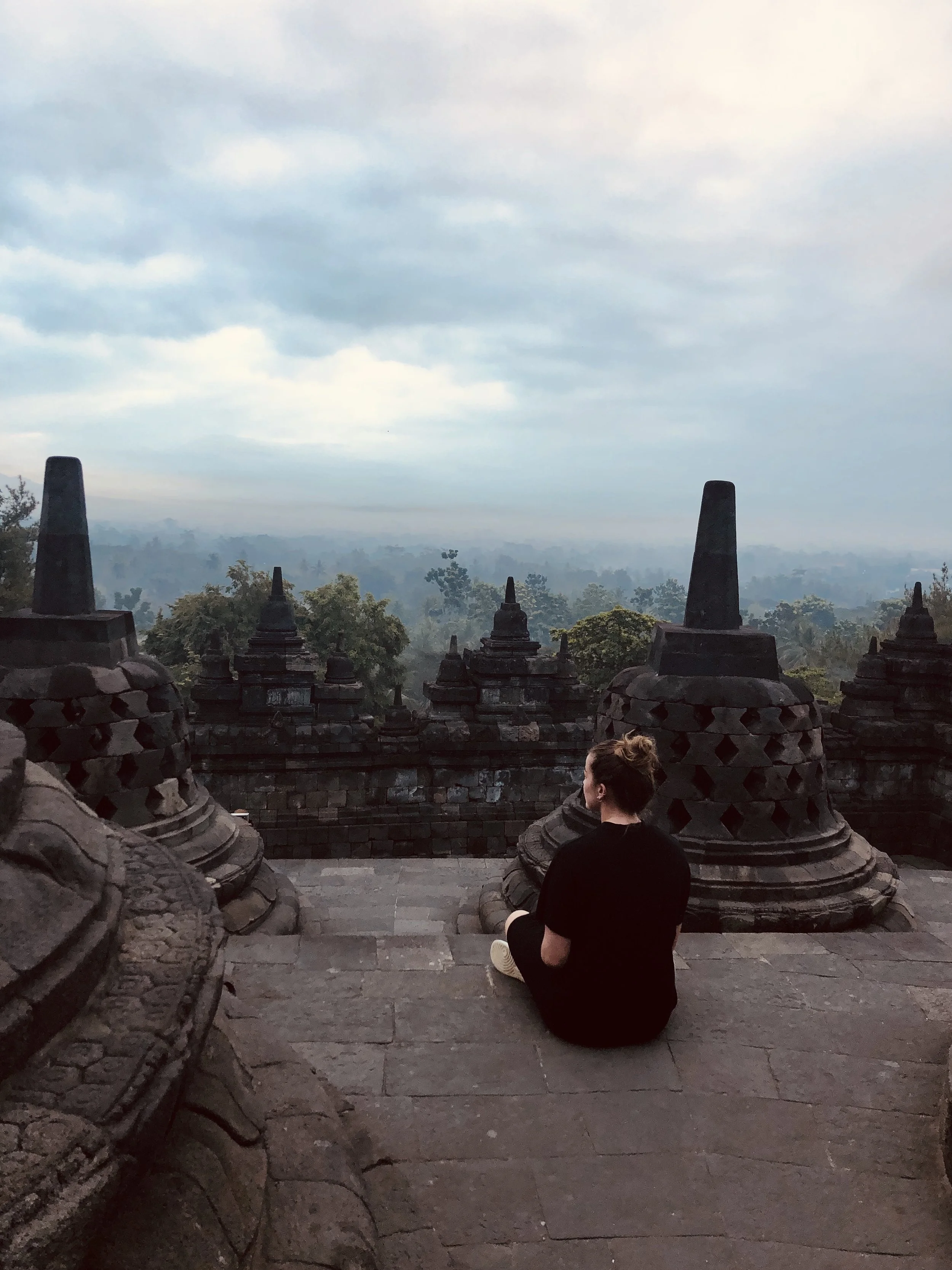 Een persoon zittend op trappen tussen oude stenen bouwwerken met een uitzicht over een uitgestrekt bos en een blauwe, bewolkte hemel.