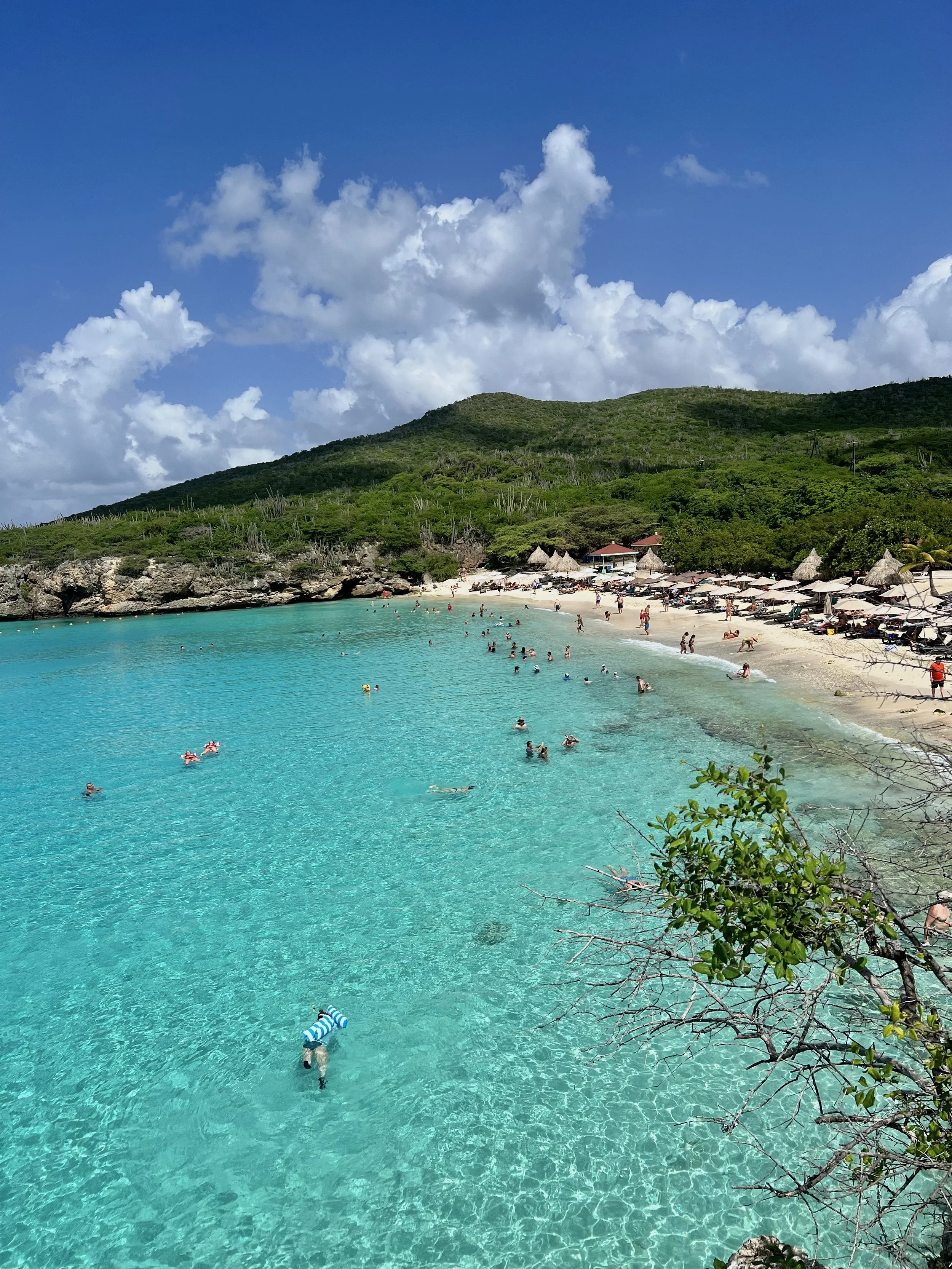 Een tropisch strand met helderblauw water, zwemmers, een zandstrand met parasols, groene heuvels en een blauwe lucht met witte wolken.