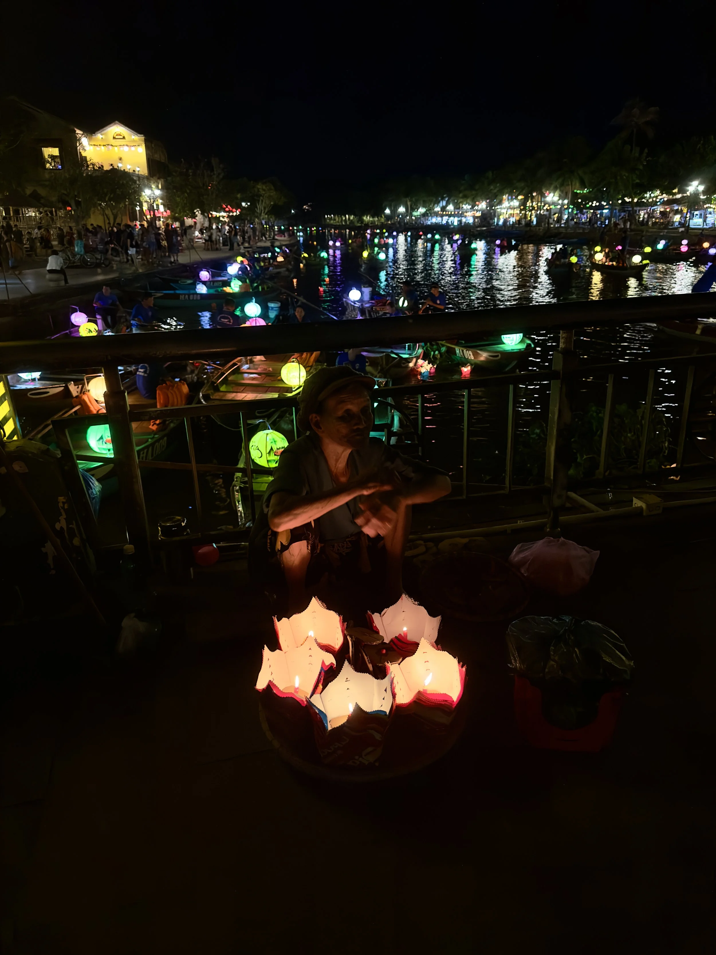 Vrouw zit op de grond omringd door brandende lantaarns. Op de achtergrond een verlichte kade met boten en mensen, bij nacht.