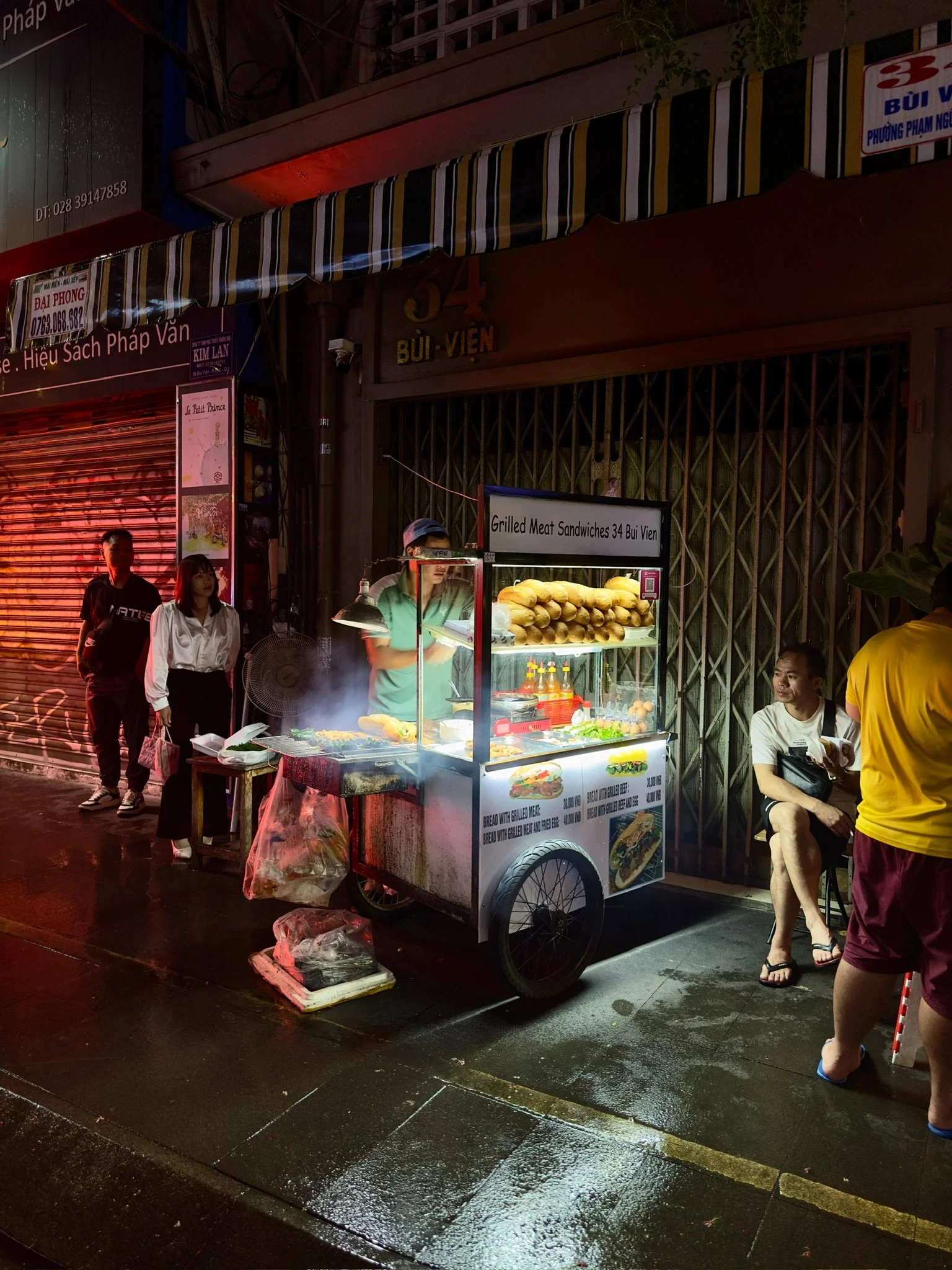 Straatkraam verkoopt gegrilde vleesboterhammen op een wagentje, omringd door mensen op een natte straat in de avond.